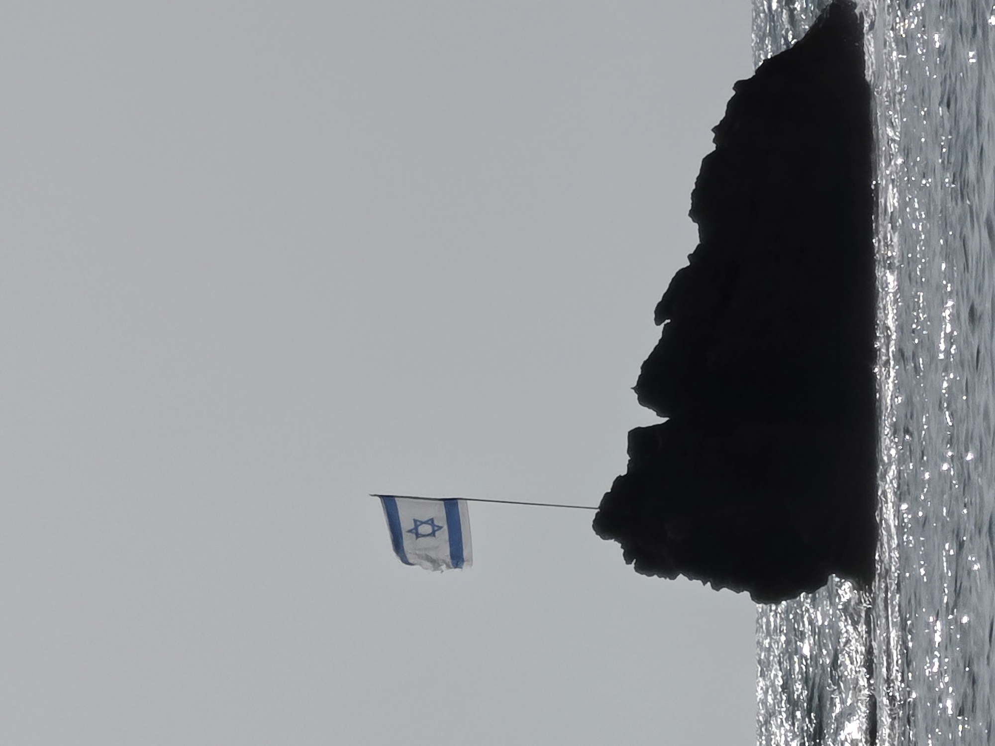 The image captures a coastal scene in Bat Yam, Israel, featuring an Israeli flag, a dark rocky outcrop, and sparkling water under a bright sky.
The central subject is a white Israeli flag, adorned with two horizontal blue stripes and a blue Star of David in the center. The flag is attached to a thin pole, extending from a rugged, dark landmass. It appears slightly wind-blown or tattered, indicating natural elements at play.
The rocky outcrop, prominent on the right side of the frame, is largely silhouetted in deep black, suggesting it is viewed against a brighter light source. Its irregular contours hint at a cliff face or a large rock formation extending into the sea.
To the right of the rocky mass, the water's surface is highly reflective, shimmering with countless bright glints of light, indicating sunlight reflecting off small waves or ripples. This strong reflection suggests it is daytime. The sky, occupying the upper and left parts of the image, is a uniform light grey, possibly overcast or hazy, providing a bright, somewhat diffused background for the flag and rock.
No specific human or animal activity is depicted; the scene is a static landscape shot. There is no discernible text beyond the Star of David on the flag. The overall impression is one of a serene but exposed coastal environment, with the national flag standing prominently against the sky and sea.