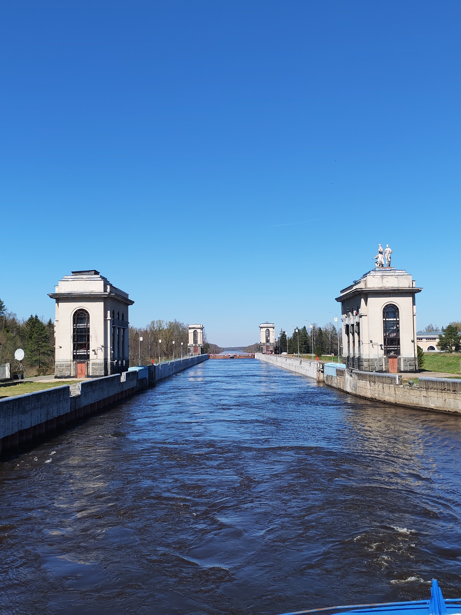 The image shows the Volga-Don Shipping Canal in Russia, specifically the structures at Lock No. 14 in Tempy. The canal stretches into the distance, flanked by concrete walls. On either side of the canal are buildings with arched windows and classical architectural details. The building on the right has statues on the roof. Trees and some grassy areas are visible on the canal's banks. The sky is clear and blue. The water is dark and turbulent.