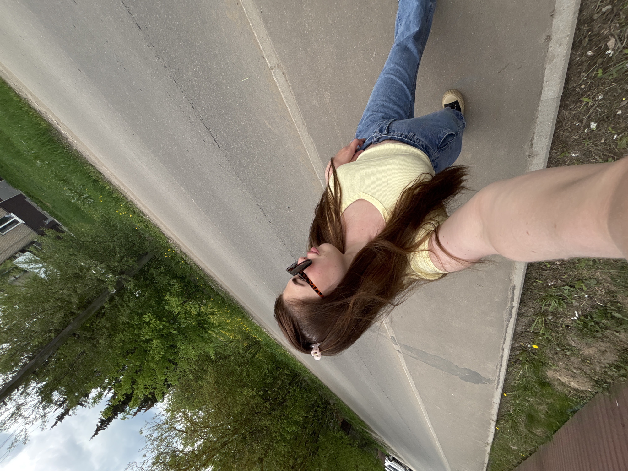 The image shows a woman in Poсёлок комбината стройматериалов-1, Russia, taking a selfie outdoors. She is wearing a yellow tank top, blue jeans, and sneakers, and has sunglasses on her head. She is standing on a sidewalk next to a road. The background includes trees and a building.