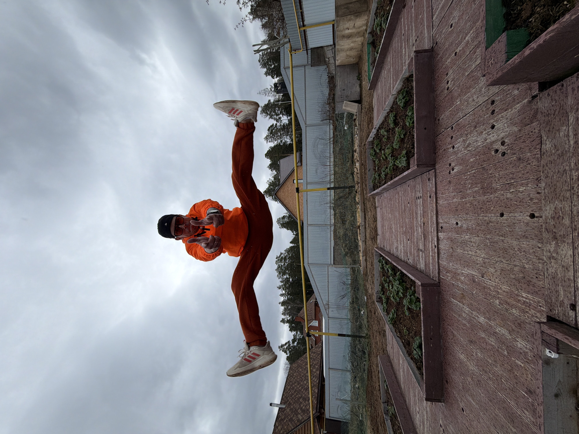 An outdoor image captures a person mid-air against an overcast sky, oriented 90 degrees clockwise from a standard upright perspective. The central subject, likely a male, wears an orange hooded jacket, dark orange pants, a black beanie, and sunglasses. They are wearing white sneakers with distinctive red stripes, characteristic of the Adidas brand. The person is performing a dynamic jump or kick, with one leg raised high. Both hands are held up, making "V-signs" or peace gestures towards the camera, and a silver ring is visible on one finger.
To the right of the person, a series of dark brown wooden structures resembling tiered raised garden beds or planters are visible. These planters contain dark soil and small green plants. The background reveals a somewhat sparse landscape under a cloudy, grey sky. Visible elements include pine trees, the roofline of a residential house, and a fence composed of both chain-link and corrugated metal. A yellow gas pipeline stretches across the middle ground. On a concrete block near the fence, faint text "02 12 18" and "2 018" is discernible.
The scene appears to be a backyard or garden area, possibly in a rural or suburban setting like Chaikovsky, Russia. The cloudy weather indicates a lack of direct sunlight. The person's pose and gesture convey a sense of playfulness and energy.