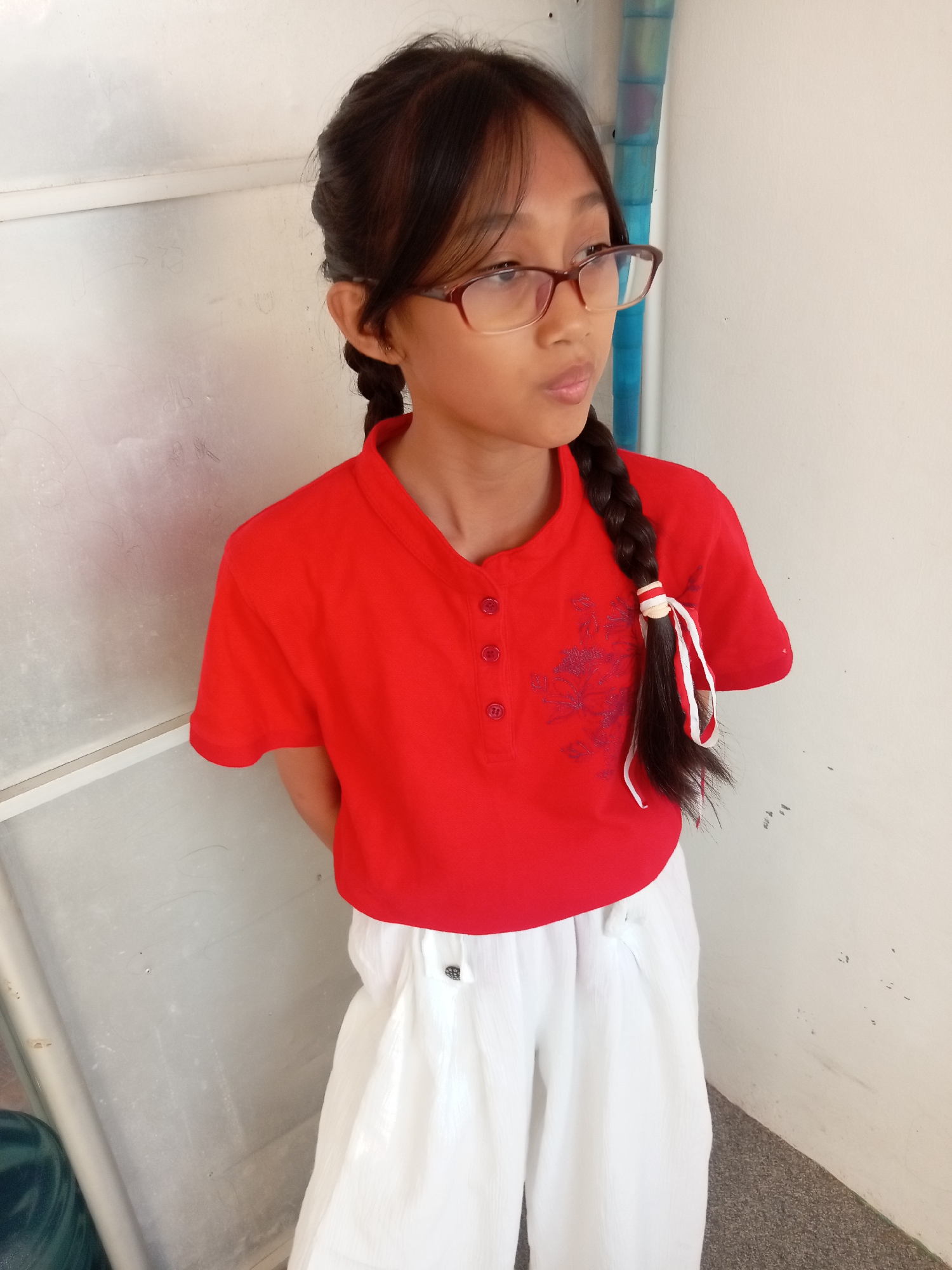 The image shows a young girl with dark hair styled in two braids, wearing glasses, a red polo shirt, and white wide-leg pants. She is standing with her back to a textured white wall, with a vertical element of blue and silver pipes visible to her right. Her hands are clasped behind her back, and she is looking to her right. The lighting suggests it is daytime, possibly outdoors or in a well-lit indoor space. The environment appears to be a simple, possibly institutional or residential, setting. The image does not provide specific landmarks or context to definitively place it within the city of Batam, Indonesia.