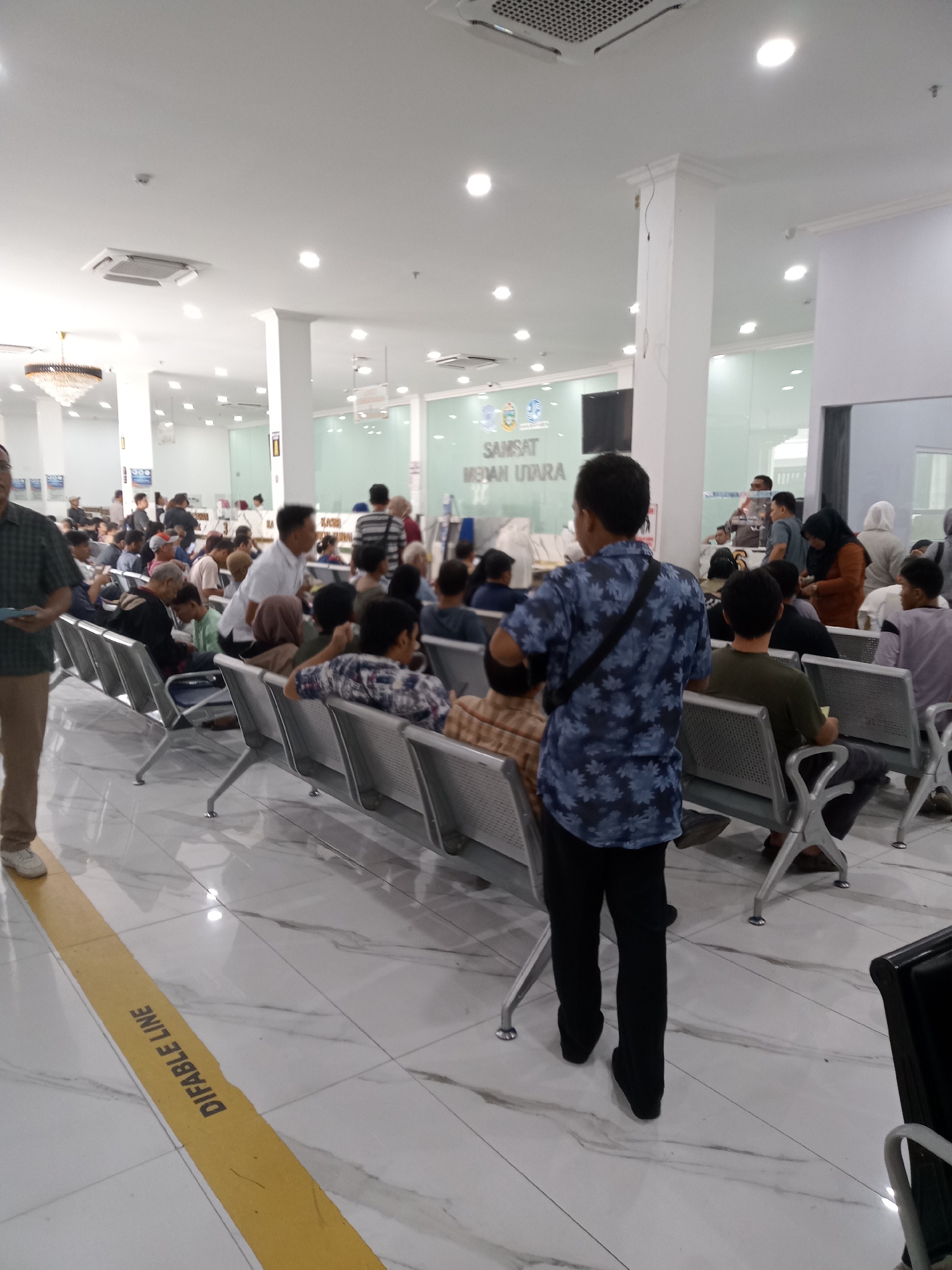 This image captures the interior of a government office, likely a Samsat (motor vehicle tax office) in the city of Medan, Indonesia. The scene depicts a waiting area filled with numerous individuals, predominantly men, seated on rows of metallic chairs. The environment is well-lit by overhead fluorescent lights, with a tiled floor and white walls.
Several people are visible in the foreground and midground. On the left, a man in a checkered shirt and beige pants stands facing away from the camera, holding a blue object. Behind him, a group of people are seated, some conversing. In the center of the image, a man in a blue floral shirt and black pants is standing, looking towards the seated individuals. Further back, more people are waiting, with some standing and others seated. A sign on the wall in the background reads "SAMSAT MEDAN UTARA," confirming the location. The overall atmosphere suggests a busy public service facility.
There's a yellow line on the floor with the words "DIFABLE LINE" printed on it, indicating a designated path for individuals with disabilities. The presence of multiple individuals waiting suggests that services are being provided, and the general arrangement of the space points to an organized, albeit crowded, administrative building.