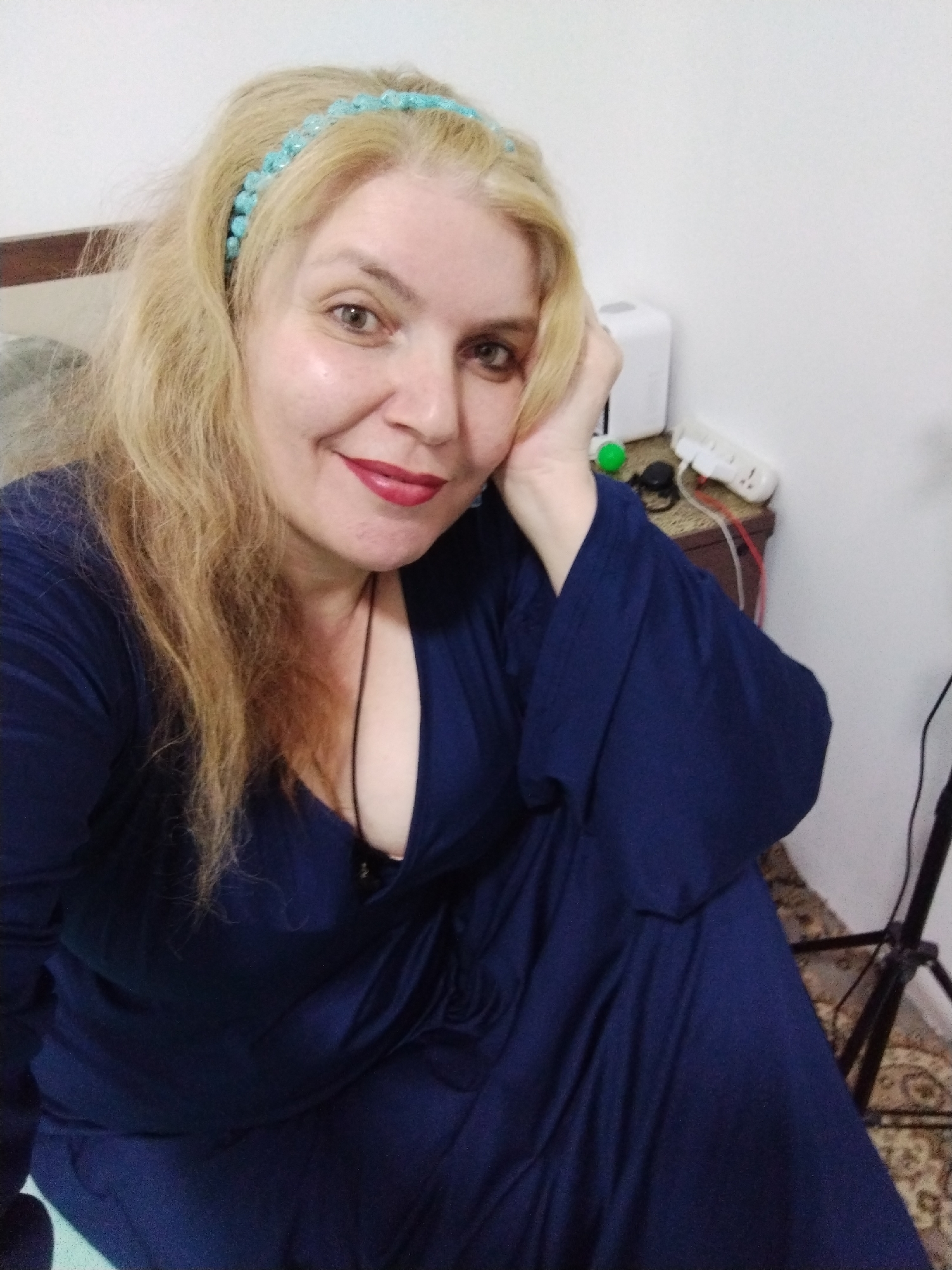 A woman with blonde hair and a blue headband sits in a dark blue dress. She rests her head on her left hand. A nightstand is behind her with a humidifier, electrical plug, and other miscellaneous objects. A tripod stand is visible in the lower right. The location is implied to be in Al Mas'oudi, United Arab Emirates.