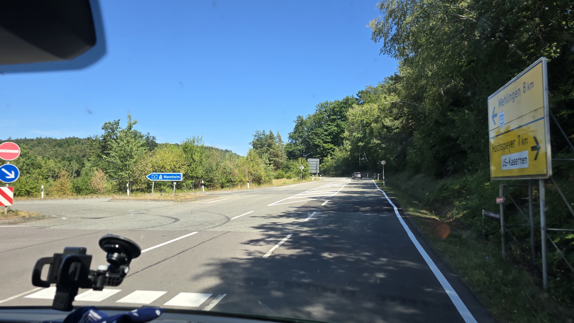 The image shows a view from a vehicle driving on a sunny day. The scene is a road intersection with trees on either side and a blue sky overhead. There are several road signs visible. On the left, there are signs indicating "No entry" and an arrow pointing left. Further down the road, a blue sign points to "Mannheim". On the right, a yellow directional sign indicates "Mehligen 8 km" to the left and "Hochspeyer 7 km" and "US-Kasernen" to the right. The road has white lane markings. A car is visible driving in the distance on the right side of the road. The foreground shows a dashboard with a suction cup mount for a phone or GPS device. The overall environment appears to be rural or suburban.