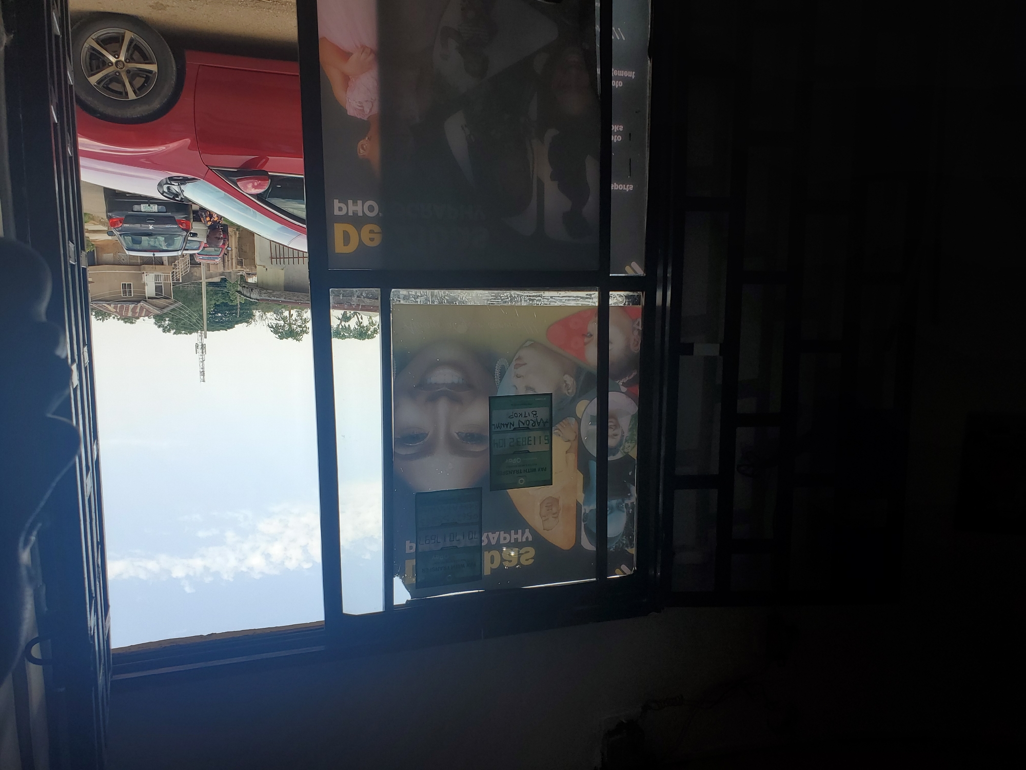 The image, taken from indoors, looks through a window displaying commercial posters, partially obscuring an outside view. The posters appear to advertise beauty products or services, with prominent facial imagery and text.
Outside, a view of what seems to be an urban setting is visible, featuring cars parked along a street, buildings, trees, and a blue sky with scattered clouds.
The image seems to be taken in Jos, Nigeria, as evidenced by the inscription "HARON NANYIL BITKOP" on one of the stickers in the window.