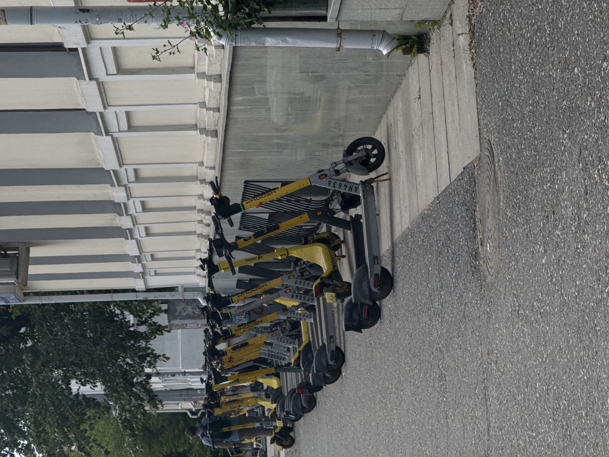 The image shows a row of yellow and gray electric scooters parked along a sidewalk in front of a building. The building has a light-colored facade with vertical gray stripes and decorative elements. A person with a backpack is walking away from the scooters on the left side of the image. The scooters are lined up neatly, and some have visible identification numbers. A downspout from the building is visible on the right, with some greenery growing around its base. The ground is paved with asphalt and concrete. The scene appears to be in an urban environment, likely a street or public area in Yekaterinburg, Russia, given the presence of the "WHOOSH" branded scooters, a popular rental service in Russian cities.