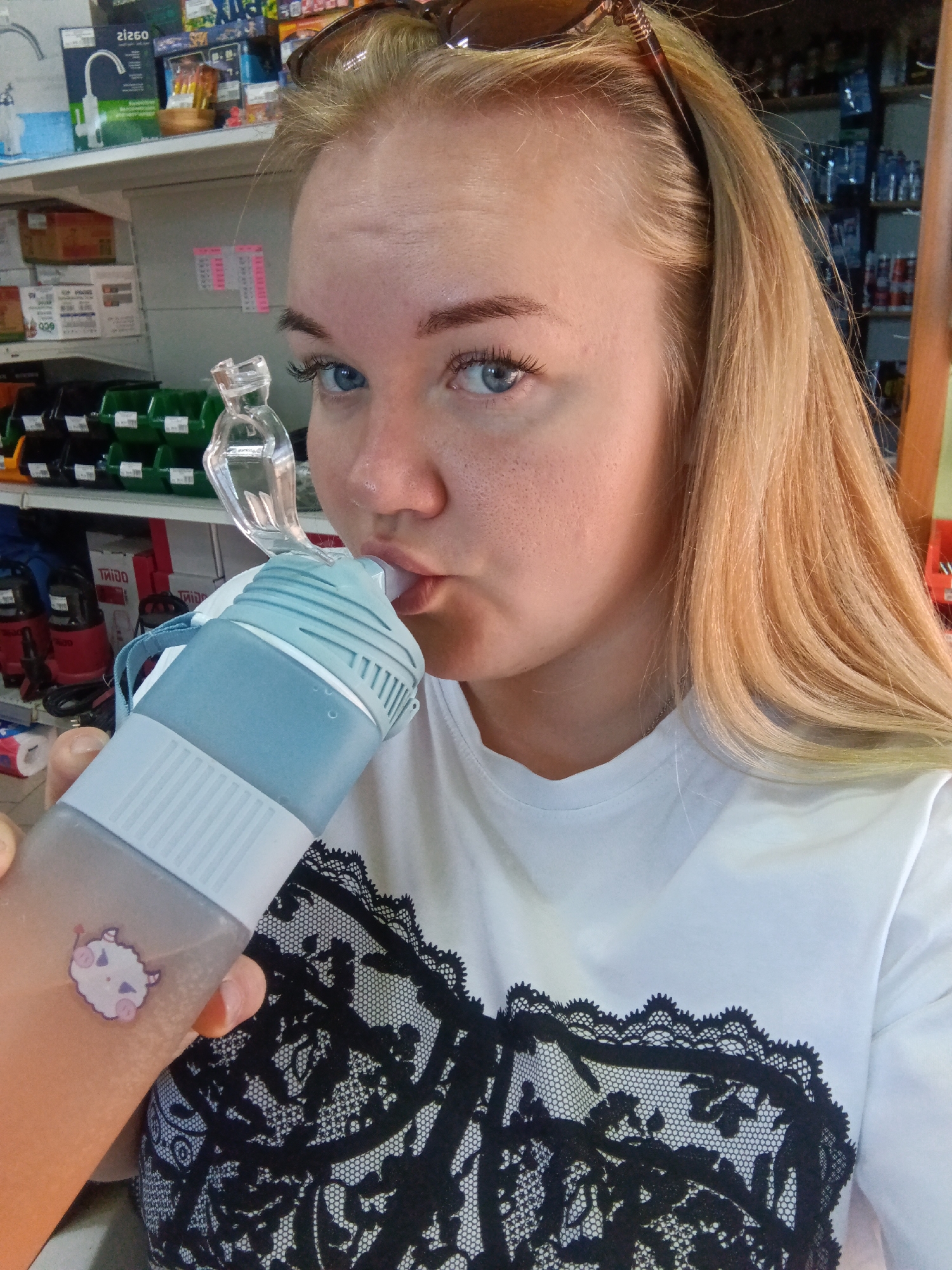 A young woman with blonde hair and blue eyes is taking a selfie while drinking from a blue and white water bottle with a clear plastic straw. She is wearing a white t-shirt with a black lace pattern on the front. The background appears to be an indoor store with shelves stocked with various items. The image does not provide any specific details or context that would allow for an identification of the location within the city of Фоки, Russia.