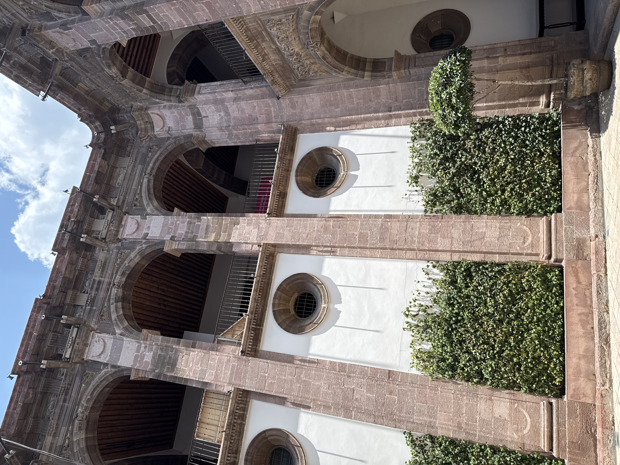 Here is a detailed description of the image:
The image depicts a vertical, upward-angled view of a multi-story building facade in San Miguel de Allende, Mexico. The building is constructed with a mix of light-colored stone, likely sandstone, and white-painted walls. The architectural style includes arched openings, some with railings, revealing interior spaces with wood beams. There are round windows with grilles set into the white wall sections. Planter boxes covered in lush green foliage vertically line the building's exterior. The lighting suggests it is daytime with clear shadows. The sky is partially visible in the upper left corner with a few clouds. There is no visible people in the image.