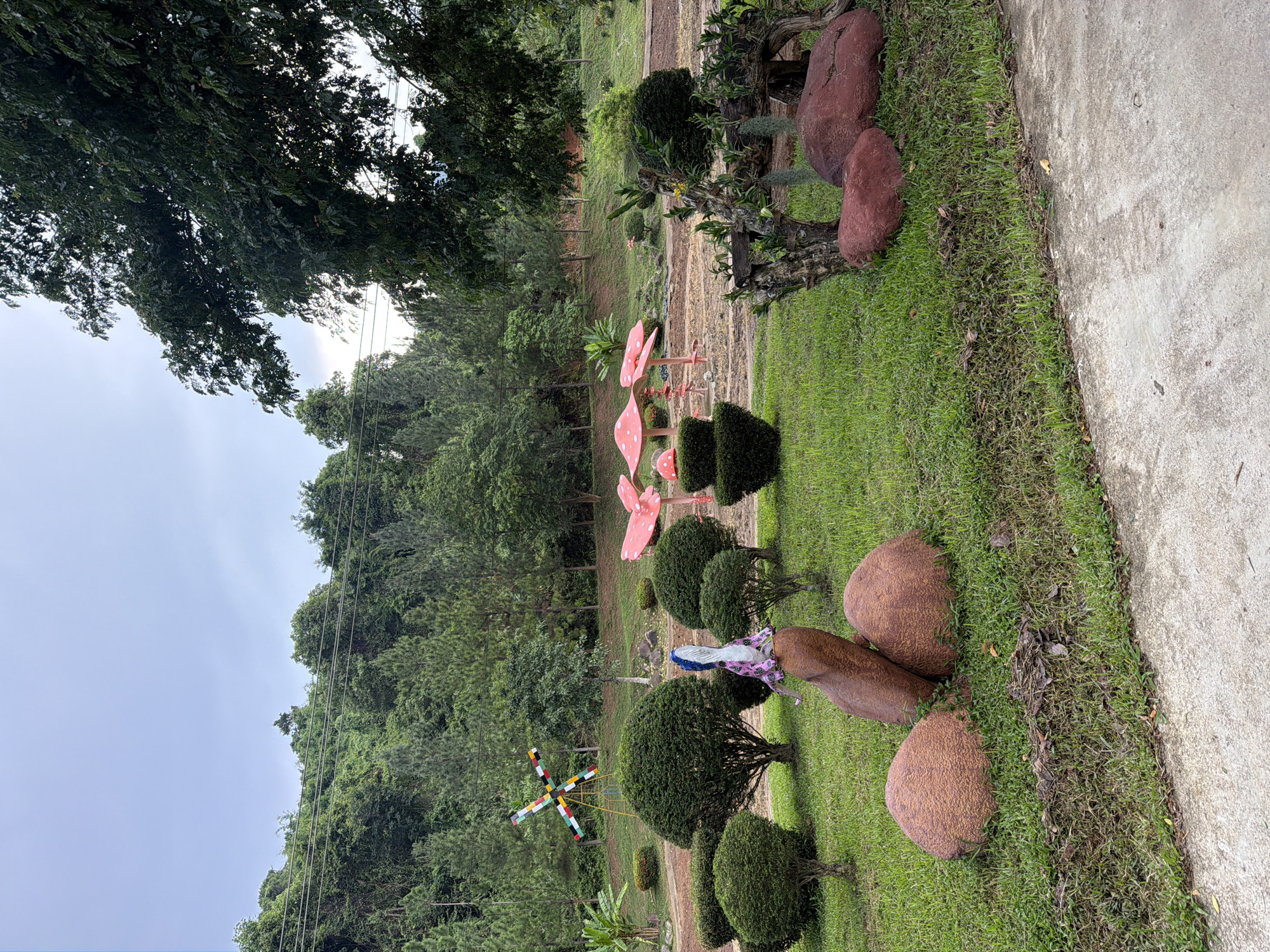 The image depicts an outdoor park area with lush green grass, manicured bushes, and a hill covered in trees in the background. Several decorative elements are visible: large, reddish-brown, rounded rocks are placed on the grass, with one large, upright stone in the center. Atop this upright stone sits a figure draped in a pink and blue patterned fabric, resembling a doll or statue. To the left of the central rock formation, a colorful, pinwheel-like structure is positioned. In the mid-ground, there are several large, pink mushroom-shaped decorations with white spots. Further back, a row of short, rounded green bushes lines a path. The environment suggests a park or garden setting, possibly in Khánh Vĩnh, Vietnam, given the tropical foliage and vegetation. The sky is overcast, indicating a cloudy day.