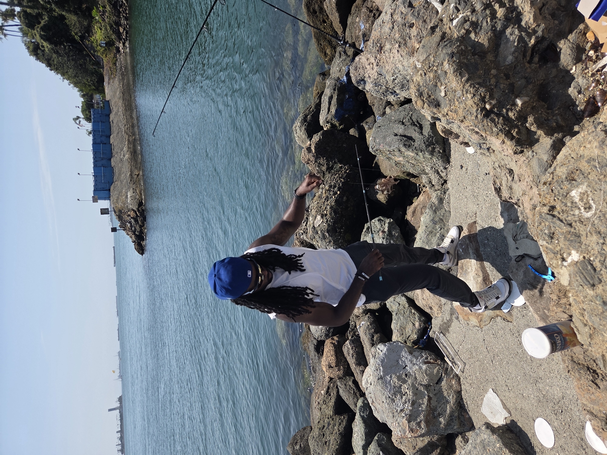 A person with dreadlocks, wearing a blue baseball cap, sunglasses, a white sleeveless shirt, and dark pants, is standing on a rocky shore, fishing with two fishing rods. The person is looking down at the fishing line. The water is calm and blue. In the background, there are several ships and a blue structure on a rocky point. The scene appears to be in Long Beach, California, judging by the style of the ships and the presence of palm trees on the distant hill. There is a can of "ANKO" brand food on the ground near the person.