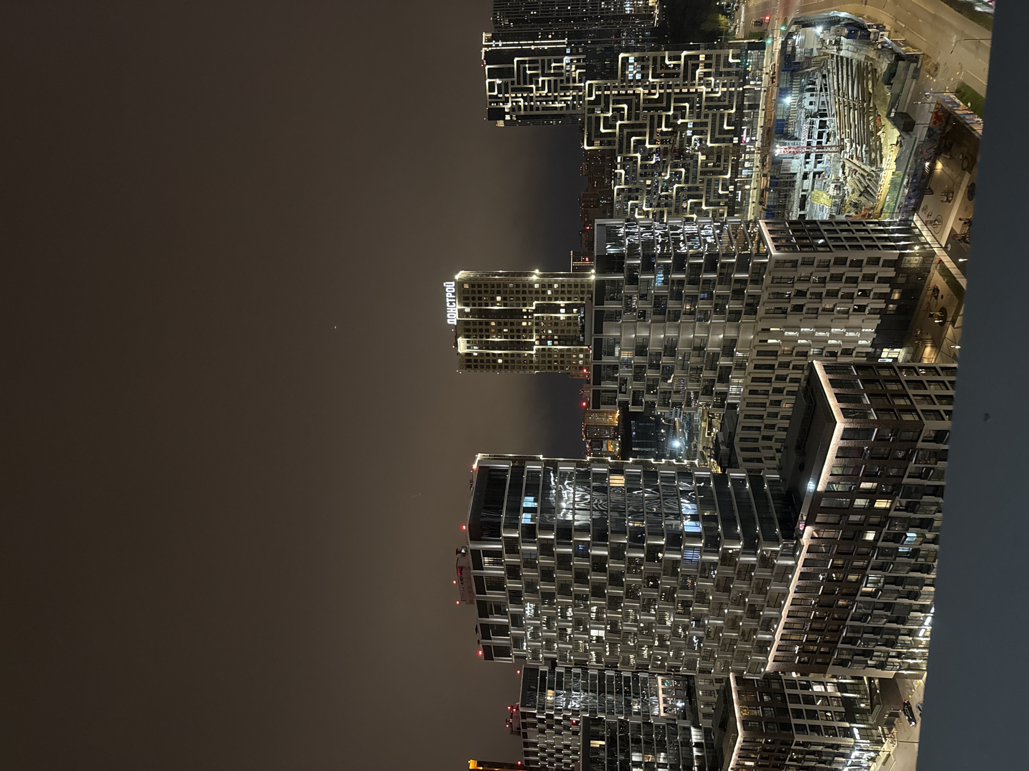 The image presents a nighttime cityscape in Khimki, Russia, captured from an elevated perspective. The scene is dominated by numerous modern, high-rise residential or commercial buildings. The dark sky indicates late night or early morning, with clear visibility.
The architecture of the buildings is distinctive, featuring numerous illuminated windows and intricate patterns of exterior lights that highlight their contemporary designs. One prominent building in the mid-ground displays a large, illuminated sign with the Cyrillic text "ДОНСТРОЙ" (DONSTROY), a well-known Russian development company.
To the right side of the image, a large construction site is brightly lit, showcasing active development. Scaffolding, various building materials, and at least one tower crane are visible, indicating ongoing construction work. Beyond the construction, illuminated roads and what appear to be vehicle lights can be seen. The overall impression is that of a bustling and expanding urban environment. No individuals are explicitly visible, but the extensive lighting and ongoing construction imply human activity within the city. The artificial lights from the buildings and construction site create a vibrant glow against the dark sky.