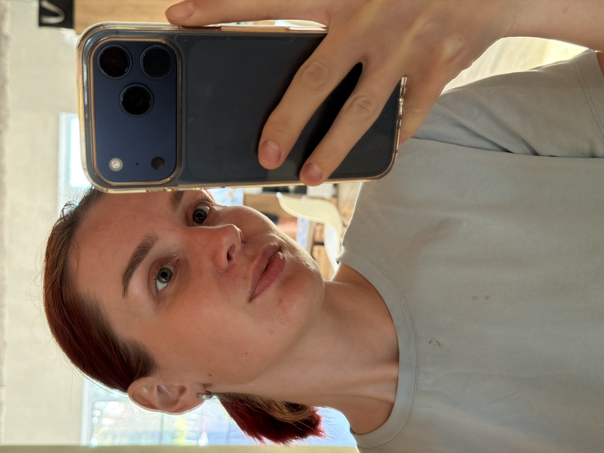 A light-skinned person is taking a selfie indoors, holding a blue and gold smartphone. The person has reddish-brown hair and light green eyes. The person is wearing a light gray t-shirt with a few stains. The location is not evident from the image. The city of Chaikovsky, Russia, cannot be determined.