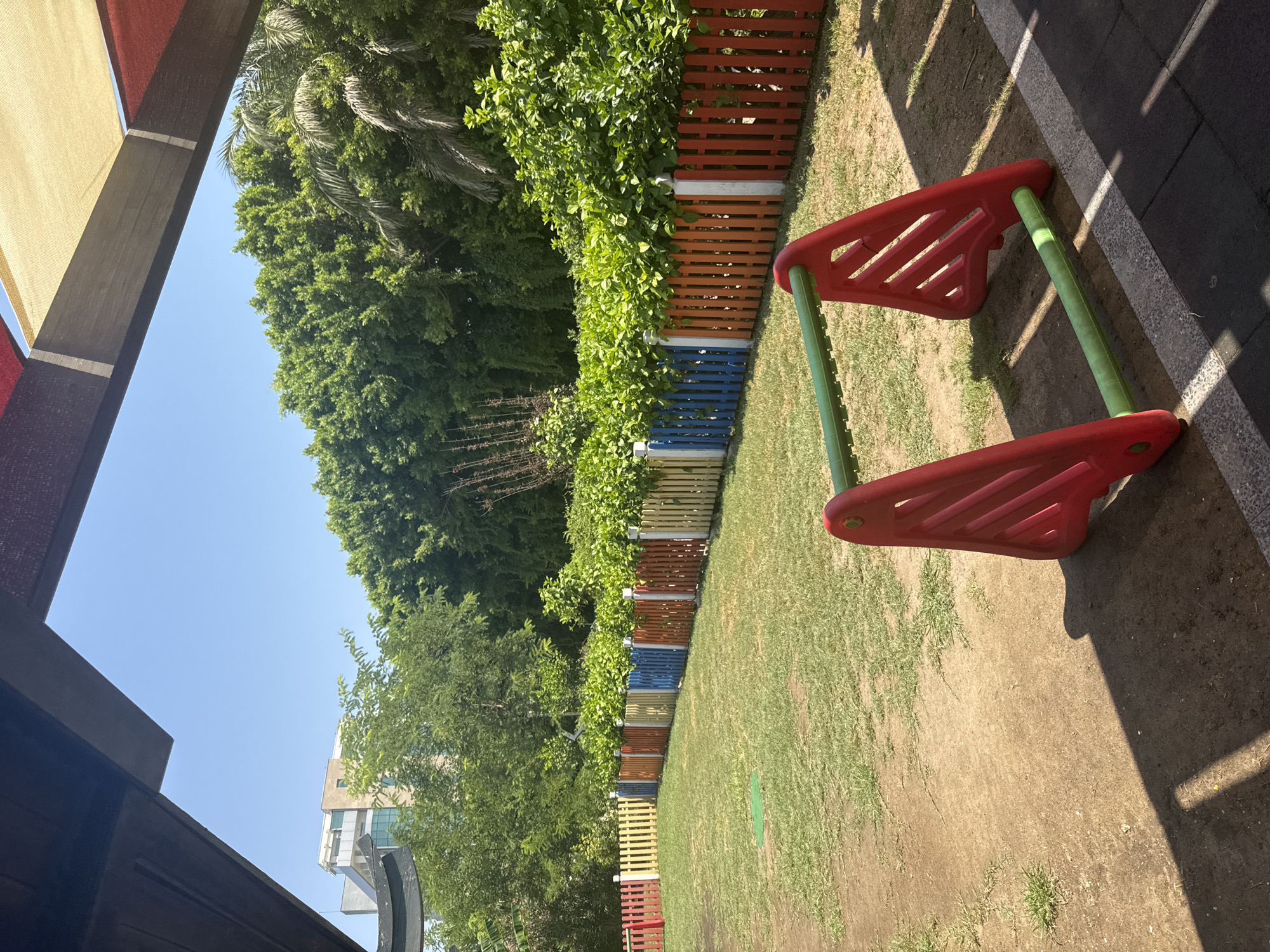 The image shows an outdoor playground scene. In the foreground, a red and green plastic A-frame structure, possibly a soccer goal or part of a climbing toy, is visible. The ground is a mixture of dirt and patchy grass. A colorful, low picket fence, composed of alternating red, orange, blue, yellow, and white posts, stretches across the middle ground, separating the play area from a dense line of green trees and foliage behind it. In the far background, a modern, multi-story building with a light-colored facade and windows can be seen peeking above the trees. The sky is clear and bright blue. The scene is illuminated by bright sunlight, casting distinct shadows on the ground. The image was taken in Aksu, Turkey.