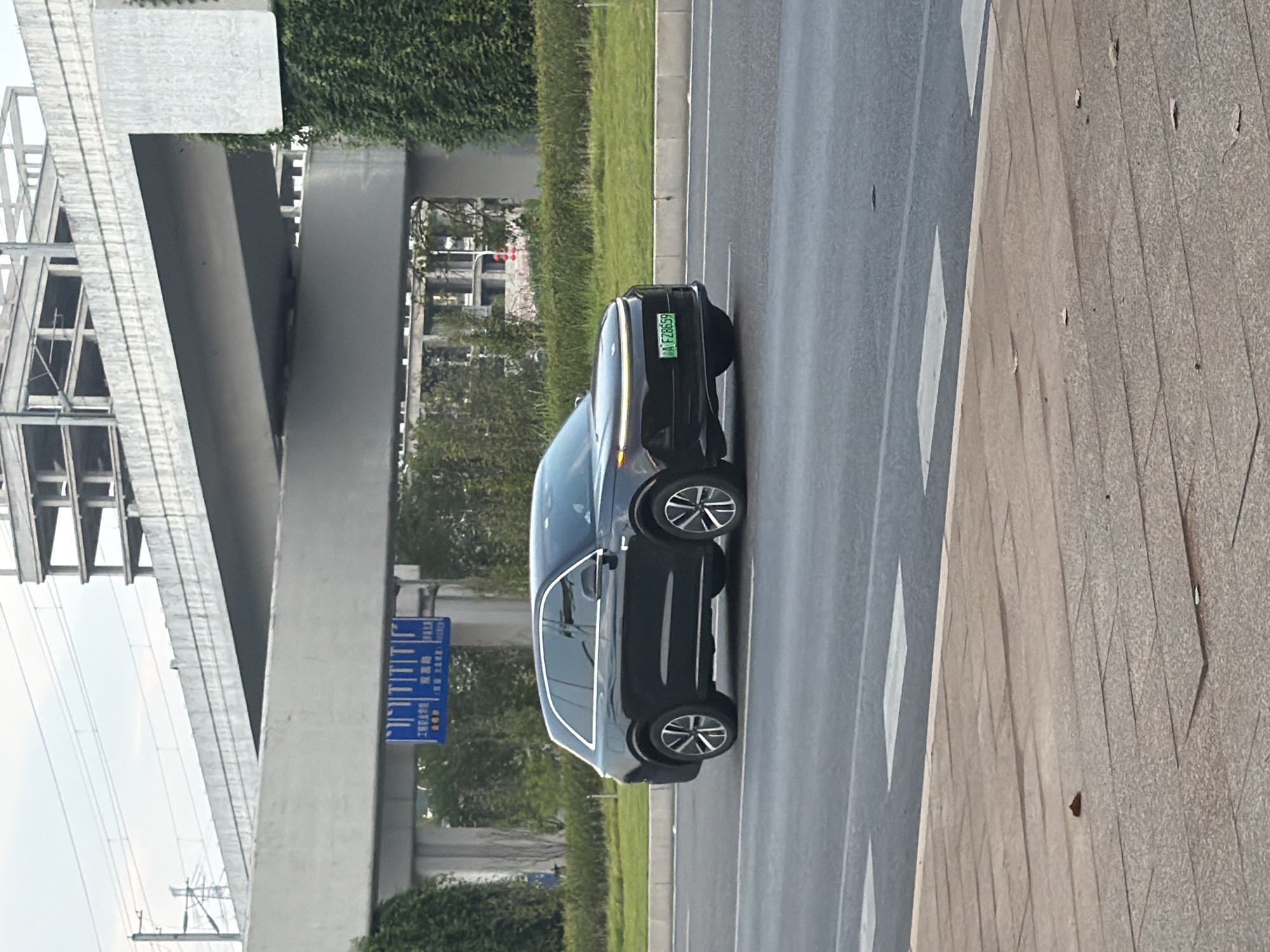 A black SUV is driving on a road in Jiangjin, China. The car is a luxury electric vehicle with a distinctive LED light bar across the front. The license plate is green with white text. In the background, an elevated highway is under construction, with scaffolding and concrete structures visible. Lush green trees and bushes line the side of the road. The sky is overcast. The image is taken from a slightly low angle, emphasizing the car and the elevated structures.