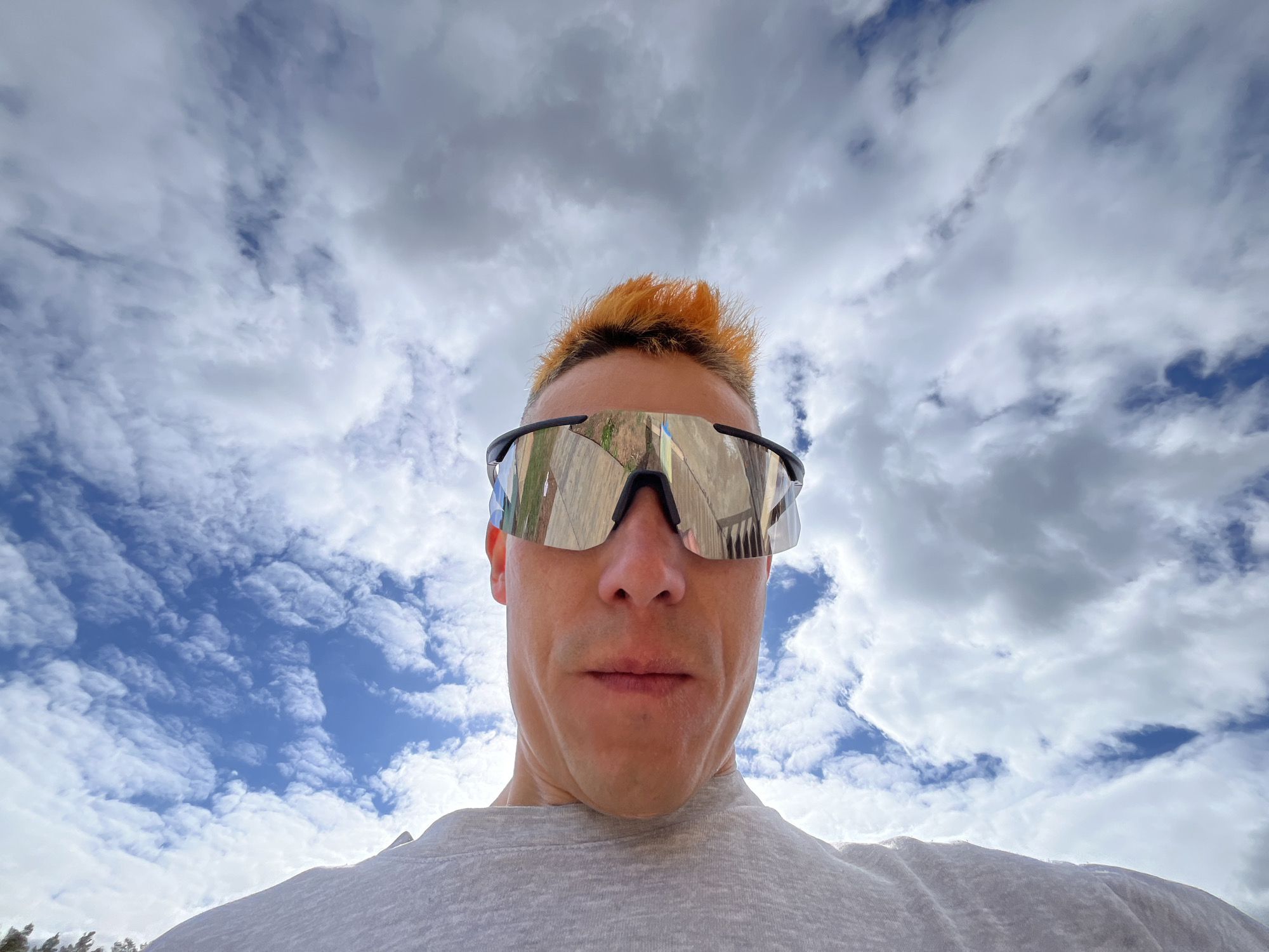 A low-angle, close-up shot shows a person wearing reflective sunglasses with a black frame and bright orange hair, facing downwards towards the camera. The person is wearing a grey t-shirt. The background consists of a blue sky with scattered white clouds. The location is Chaikovsky, Russia.