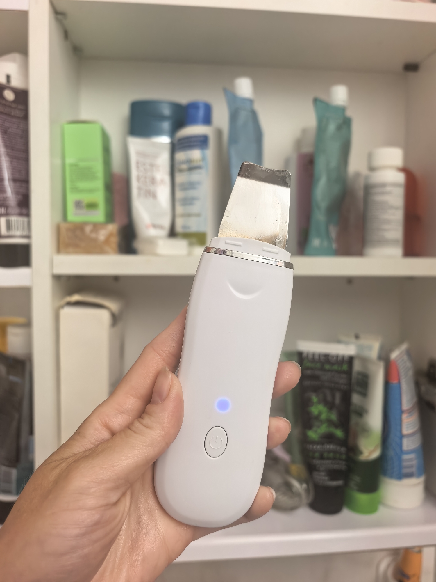 A person's hand is holding a white electronic facial cleansing device with a metal spatula attachment. The device has a power button and a blue indicator light. The background shows shelves in a cabinet filled with various toiletries and skincare products. The image appears to be taken indoors. There is no information in the image to suggest a location in Sterlitamak, Russia.