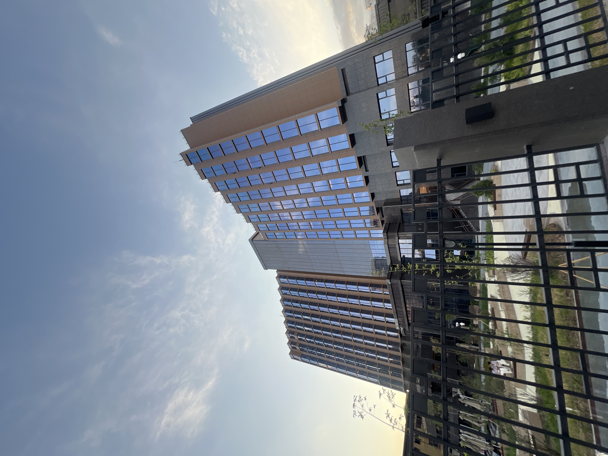 A low-angle shot captures a modern, multi-story building with a facade of brown panels and reflective blue windows, likely situated in Bishkek, Kyrgyzstan. The building's angular design is accentuated by its upward tilt in the frame. In the foreground, a black metal fence with vertical bars and horizontal supports partially obscures a landscaped area. Within this landscaped area, two figures in white attire are seated on a bench. The sky above is partly cloudy with soft, wispy formations against a pale blue backdrop, suggesting it is either early morning or late afternoon. The overall scene conveys a sense of contemporary urban architecture.