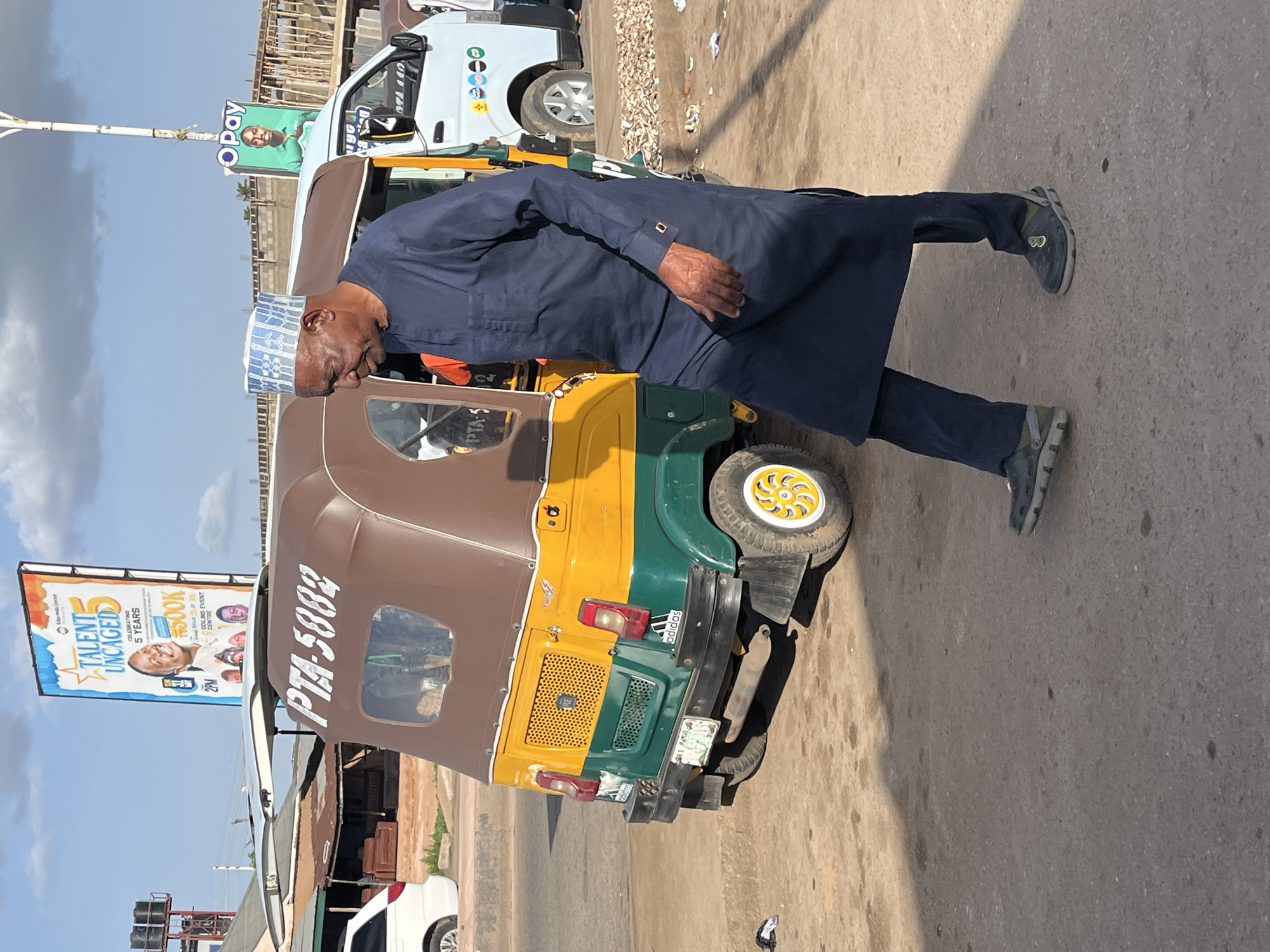 A man in a dark blue, ankle-length tunic and matching pants, wearing a blue and white patterned cap, walks across a dusty, paved road. He is looking down and to his right. Behind him, a yellow and green auto-rickshaw with a brown top is parked. The auto-rickshaw has "PTA 5882" written on its side in white letters and an "Adidas" logo on its rear bumper. To the right of the auto-rickshaw is a white van with several circular logos on its side. In the background, there are billboards, construction materials, and buildings. One billboard features a picture of a smiling person and the words "TALENT UNCAGED 5 YEARS". Another smaller billboard shows a person and the word "OPAY". The sky is blue with scattered clouds. The scene appears to be a street in Gura Top, Nigeria, likely a commercial or busy area.