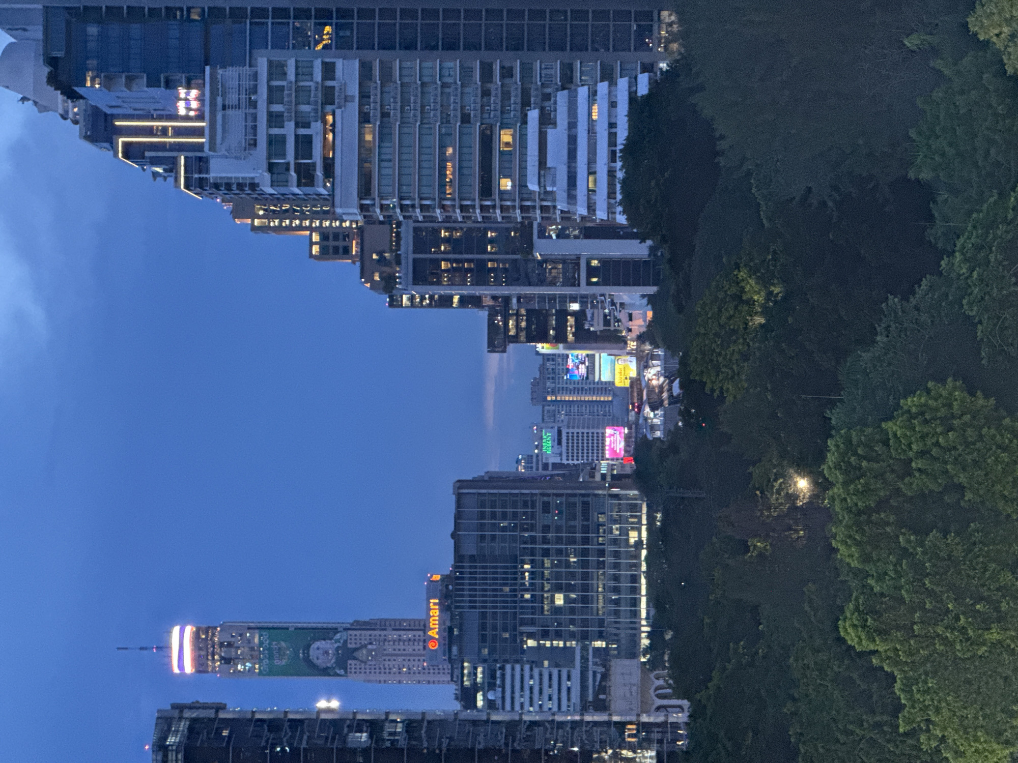 The image shows an evening cityscape in Bangkok, Thailand, featuring a mix of modern buildings and lush greenery. The perspective is slightly elevated, capturing the scene from a high vantage point.
Buildings dominate the left side, with several high-rise structures displaying varying architectural styles. Some are modern glass towers, while others have a more traditional appearance with visible balconies and window patterns. Lights from within the buildings illuminate the scene, indicating the presence of activity. The "Amari" logo is visible on one of the buildings.
To the right, dense trees and foliage are present, providing a contrast to the urban environment. The trees are illuminated by streetlights, revealing their dense canopy.
In the distance, bright lights from other buildings and streetlights add to the overall cityscape, creating a dynamic and vibrant urban landscape. The sky above is a dark blue, indicating the time of day is either dusk or early evening.