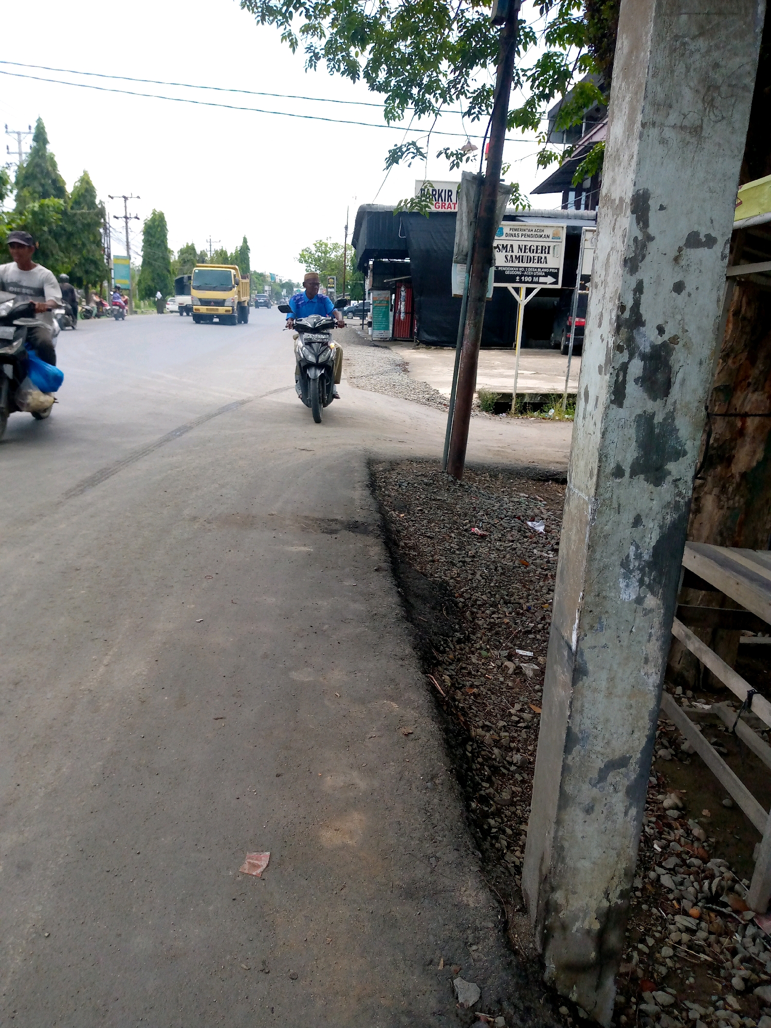 The image depicts a street scene in Keudebayu, Indonesia, with a focus on a paved road. In the foreground, to the right, is a weathered concrete pillar adjacent to a section of rocky ground and what appears to be a wooden fence or structure. The road itself shows signs of wear, including a tire mark.
Several vehicles and individuals are present on the road. A person is riding a motorcycle in the center of the frame, wearing a blue shirt and dark pants. To the left, another person is riding a motorcycle, wearing a white shirt and a dark cap, with a blue bag on the back of their motorcycle. Further down the road, a yellow dump truck is visible, along with other vehicles and motorcycles in the background, suggesting moderate traffic.
Along the right side of the road, there is a sidewalk area with a sign indicating "SMA NEGERI SAMUDERA" (Samudera State High School) and an arrow pointing in a direction, along with a distance of "2.100 MI". There are also buildings visible behind the sign. Trees line the left side of the road in the background. The sky is overcast.