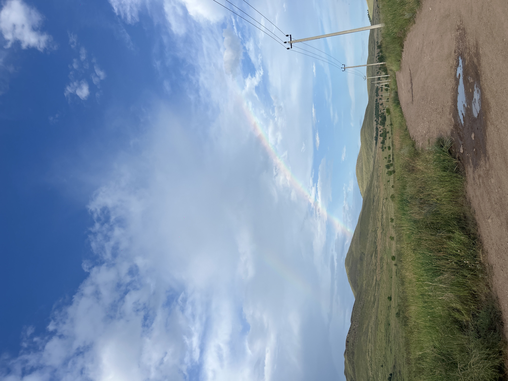 The media file depicts a natural landscape with a dirt road in the foreground, leading into a grassy hillside under a vast sky. A faint rainbow arcs across the sky, suggesting recent or ongoing rain and sunshine. The sky is a mix of bright blue and fluffy white clouds.
Along the road, several utility poles with power lines are visible, indicating a rural or semi-rural setting. The road itself appears to have some puddles, further reinforcing the idea of recent precipitation. The hillside is covered in green grass and scattered trees, with rolling hills extending into the background.
The time of day appears to be daytime, possibly late afternoon, given the angle of the sunlight and the overall brightness of the scene. The weather is dynamic, with sunshine and rain contributing to the rainbow. The location is identified as Nikolo-Petrovka, Russia, which provides geographical context. There are no people or discernible text in the image. The overall mood of the image is one of serene natural beauty, with the rainbow adding a touch of wonder.
