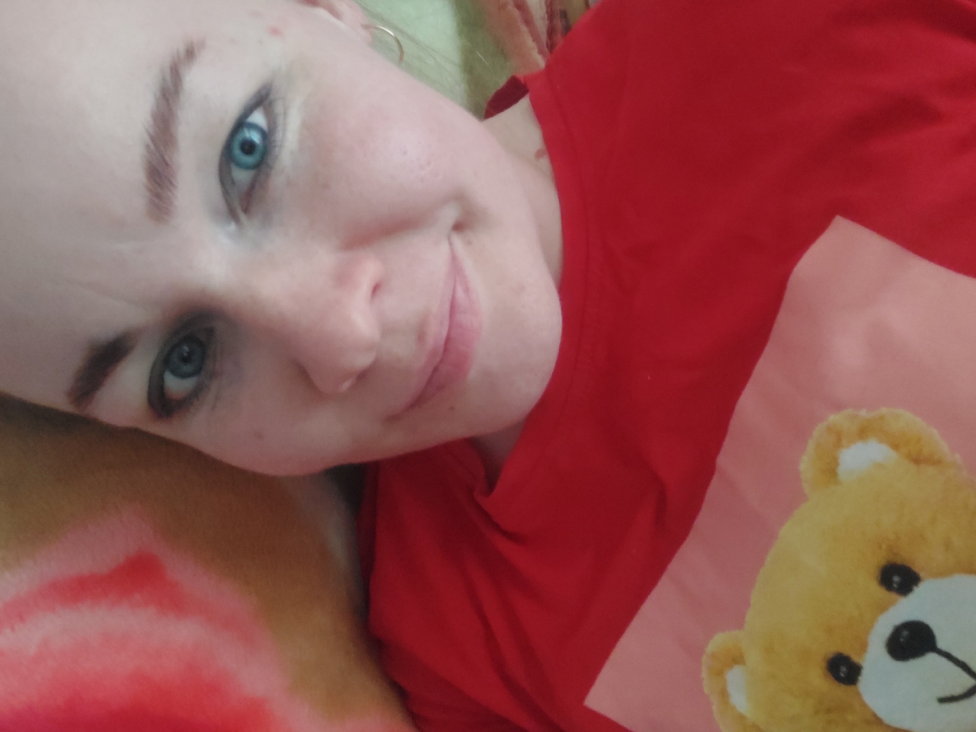 A selfie-style photograph shows a fair-skinned woman with light brown eyebrows and striking blue eyes. She is smiling slightly. A section of a red garment is visible, partially covering a pale pink surface, and a portion of a plush brown teddy bear with black eyes and a white muzzle is at the bottom right. The background is indistinct. The location is Большой Букор, Russia.