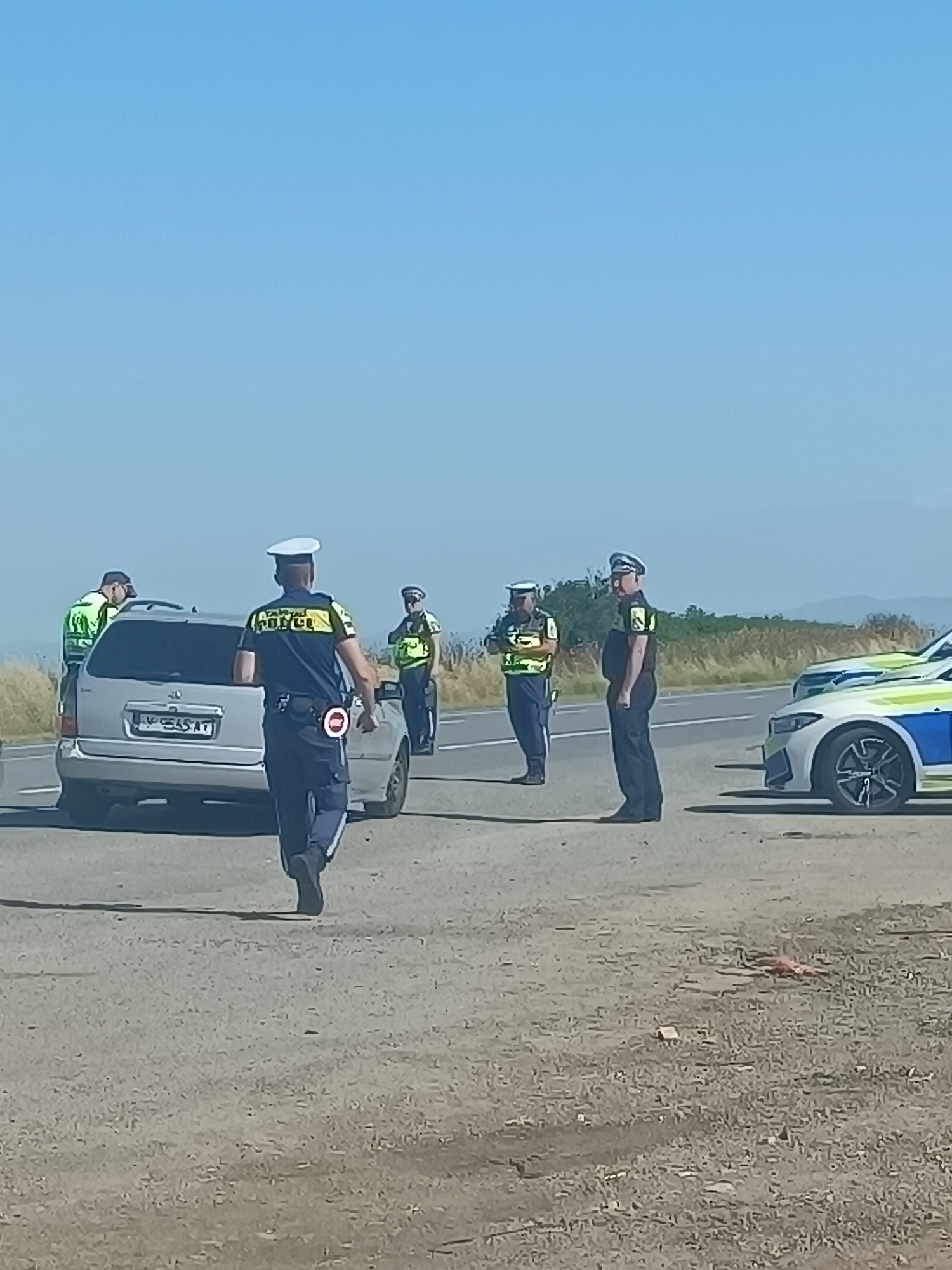 The image depicts a roadside scene in Tenevo, Bulgaria, under a clear blue sky. Several police officers in uniform are visible, some directing traffic or observing vehicles. A silver minivan is stopped on the asphalt road, with at least two officers interacting near it. Another police vehicle, a white and blue car with police markings, is parked further down the road. The surrounding environment appears to be rural, with dry grass and distant hills visible. One officer in the foreground is walking away from the camera, wearing a blue shirt with "POLICE" lettering and a white hat, and holding a red and white traffic control device.