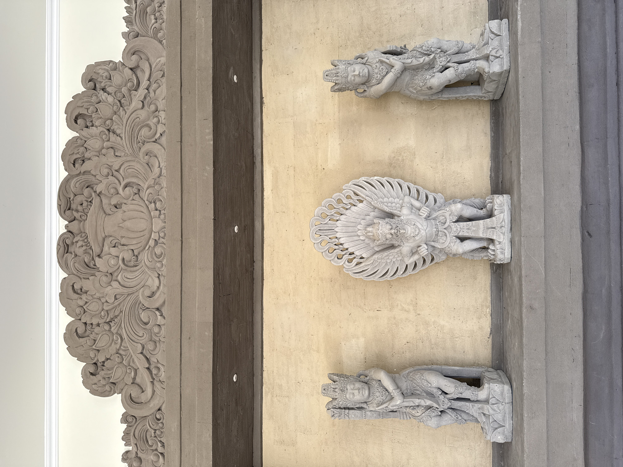 Three stone statues are displayed against a textured yellow wall, topped by an ornate stone carving. The central statue depicts a winged creature, possibly a Garuda, with outstretched wings and a fierce expression, standing on a decorative base. Flanking this central figure are two human-like statues, each wearing elaborate headdresses and traditional attire, also standing on decorated bases. The statues are positioned on a grey ledge. Above the wall, a dark brown beam runs across the width of the frame, with recessed lighting fixtures visible. The wall is framed by a pale yellow ceiling with white molding. The overall aesthetic suggests a cultural or religious setting, likely a temple or a traditional Balinese building. This scene is located in Kuta Selatan, Indonesia.