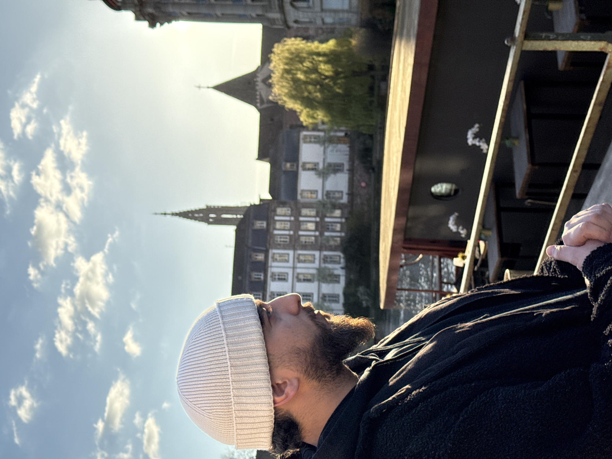 A male individual is depicted in profile, looking towards the right side of the frame, bathed in golden hour sunlight. He wears a light-colored, possibly white or off-white, ribbed knitted beanie and a dark, possibly black, textured jacket or hoodie. A prominent beard is visible on his face. His hands are loosely clasped or held together in the lower right foreground.
The scene is set in Strasbourg, France, along the Ill River. In the mid-ground, a dark-hulled riverboat or barge, likely a floating restaurant or bar, is moored, featuring several small tables with what appear to be floral decorations and a single circular porthole. A metal railing runs horizontally, separating the man from the river and the boat.
Across the river, a collection of historic buildings forms the background under a blue sky with scattered white clouds. The most prominent landmark is the distinct spire of Église Saint-Paul de Strasbourg, visible on the left side of the background. To its right, another building with a dark, pointed roof is visible, possibly another church or a part of the university complex. A vibrant green, weeping willow-type tree stands prominently on the far bank. The sunlight illuminates the entire scene, casting a warm glow, particularly on the man's beard and the water's surface. The vantage point is consistent with a location such as the Quai des Bateliers or a nearby bridge along the Ill River.