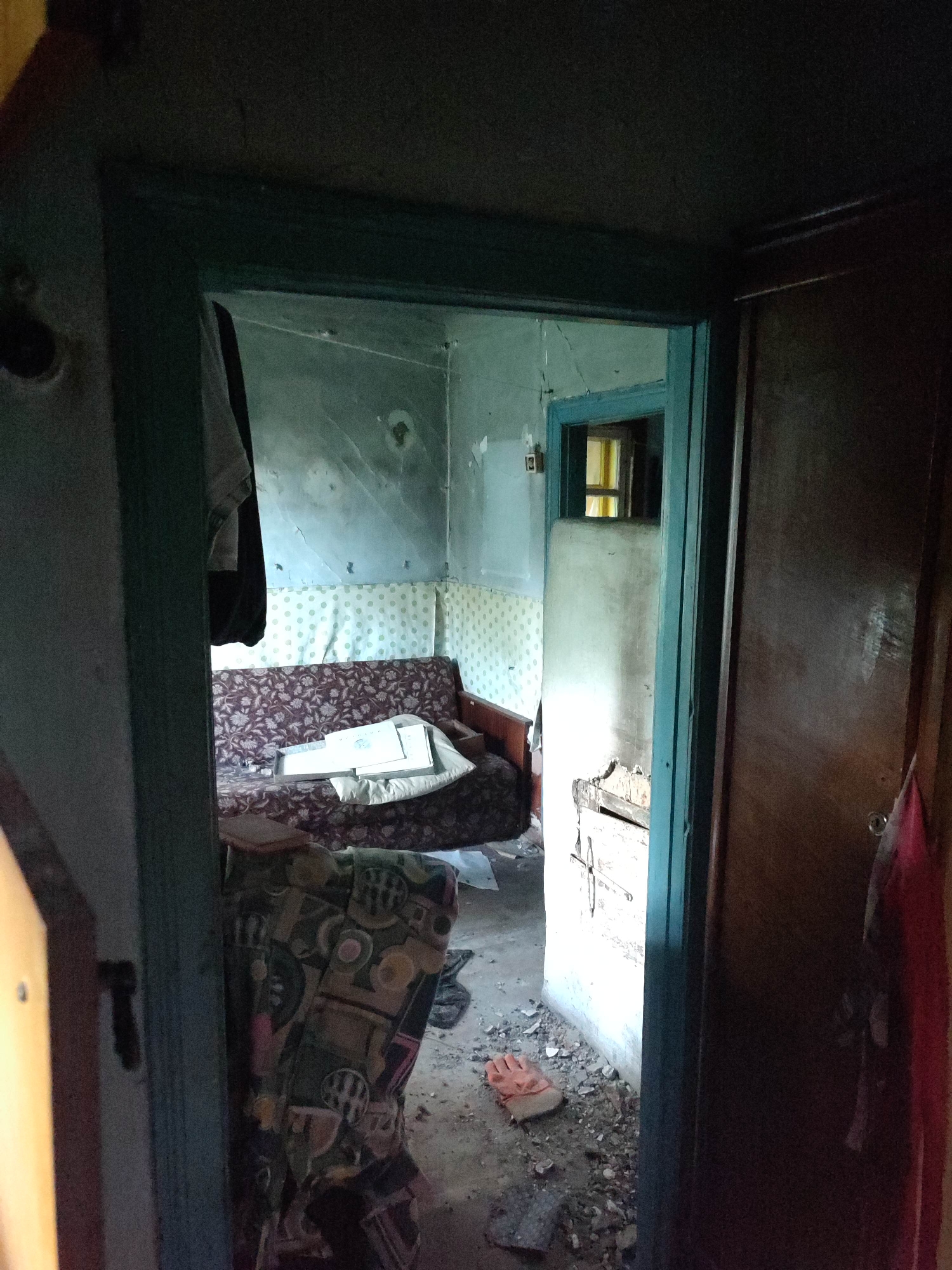 The image depicts an interior view of what appears to be a dilapidated room in Gremyachinsk, Russia. The scene is viewed from a doorway looking into another room. The interior is cluttered and shows signs of neglect.
In the foreground, to the left, is a portion of a dark wooden door frame and a sliver of a brown door. To the right, a dark wooden door is visible, with a keyhole and a hook on which a red garment is hanging.
The room beyond the doorway contains a sofa with a floral pattern, covered with papers and pillows. A patterned armchair is partially visible in the lower left of the frame. The walls are a pale color, with some patterned wallpaper visible behind the sofa. There are cracks in the walls and ceiling, and debris is scattered across the floor, including what looks like broken plaster and a single orange glove. A small window with yellow panes is visible in the back wall of the room. Clothes hang from a rod on the left side of the room, partially obscuring the wall.
The lighting suggests it is daytime, but the interior is dimly lit, indicating potential lack of natural light or a very overcast day. The overall impression is one of abandonment and disrepair. There is no visible text in the image.