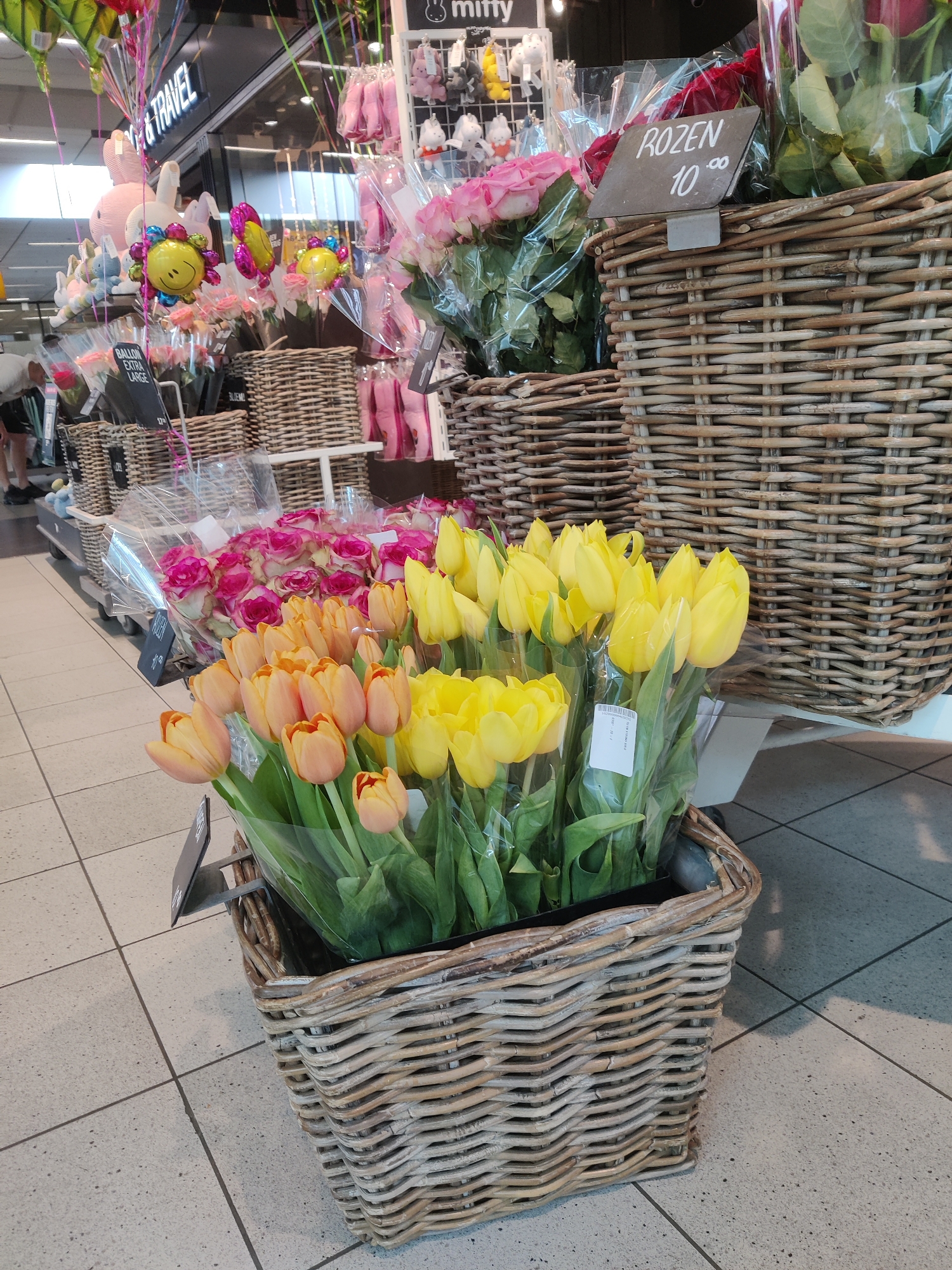 The image shows a display of flowers for sale in a retail environment at Schiphol, Netherlands. In the foreground, a large wicker basket contains an assortment of yellow and orange tulips. To the left of the tulips, a smaller wicker basket holds pink roses. Behind the flowers, there are more wicker baskets and shelves displaying various products, including balloons and stuffed animals. A sign above the roses reads "ROZEN 10.00". The floor is tiled. In the background, parts of a shop interior are visible, including signage for "TRAVEL" and "miffy". One person, wearing dark shorts and a white top, is partially visible on the far left, appearing to be walking away from the camera. The overall scene suggests a flower stall or section within a larger store, likely a supermarket or gift shop.