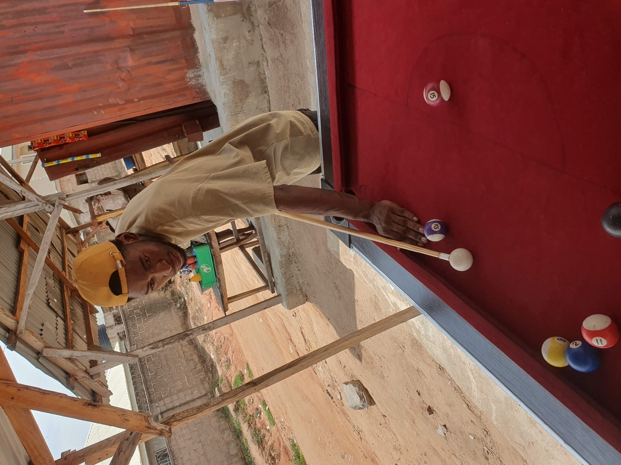 A young Black man wearing a tan t-shirt and a yellow baseball cap is playing pool outdoors in Lokoja, Nigeria. He is positioned to strike a white cue ball with a pool cue. Several billiard balls, including a yellow ball with the number 9, a blue ball with the number 2, a red ball with the number 11, a purple ball with the number 12, and a pink and white ball with the number 15, are scattered on the red felt of the pool table. In the background, there is a wooden shelter with a corrugated metal roof, a brick wall, and a dirt path. A green plastic crate containing bottles is visible on a wooden table in the background. The scene appears to be in a casual, outdoor setting.