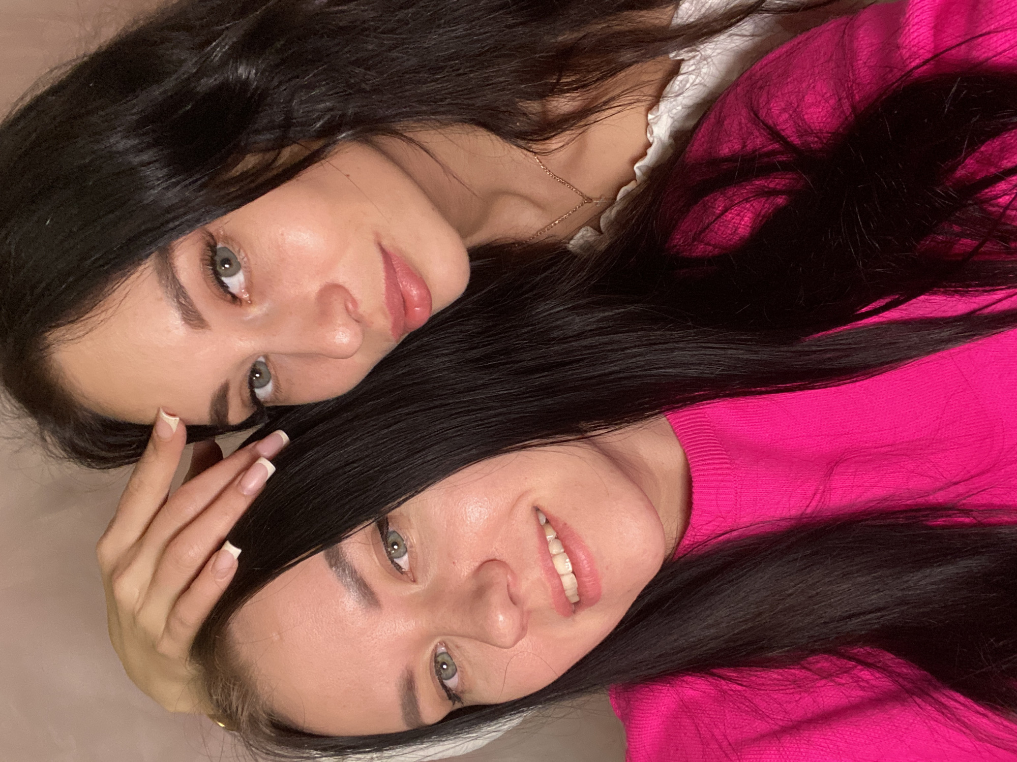 Two women with long dark hair are lying close together. The woman on top is wearing a white top with lace trim and a gold necklace. The woman on the bottom is wearing a bright pink sweater. One of the woman's hands is visible. Both women have light eyes. The background is plain and light-colored. The location is unknown, the claim of Chaikovsky, Russia is unsupported by the image.
