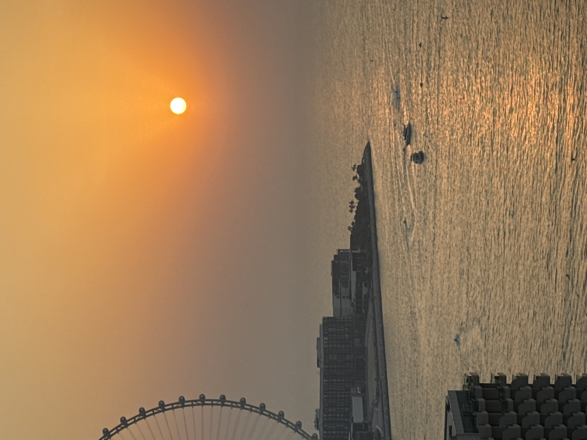 The image captures a sunset over the Arabian Gulf in Dubai, United Arab Emirates. In the foreground, the corner of a modern building with multiple balconies is visible. To the left, a portion of a large Ferris wheel, likely the Ain Dubai, is visible against the hazy orange sky. In the middle ground, a cluster of buildings forms a coastline, with several boats and jet skis scattered across the water. The setting sun casts a warm, golden glow on the ocean's surface, creating reflections and a serene atmosphere.