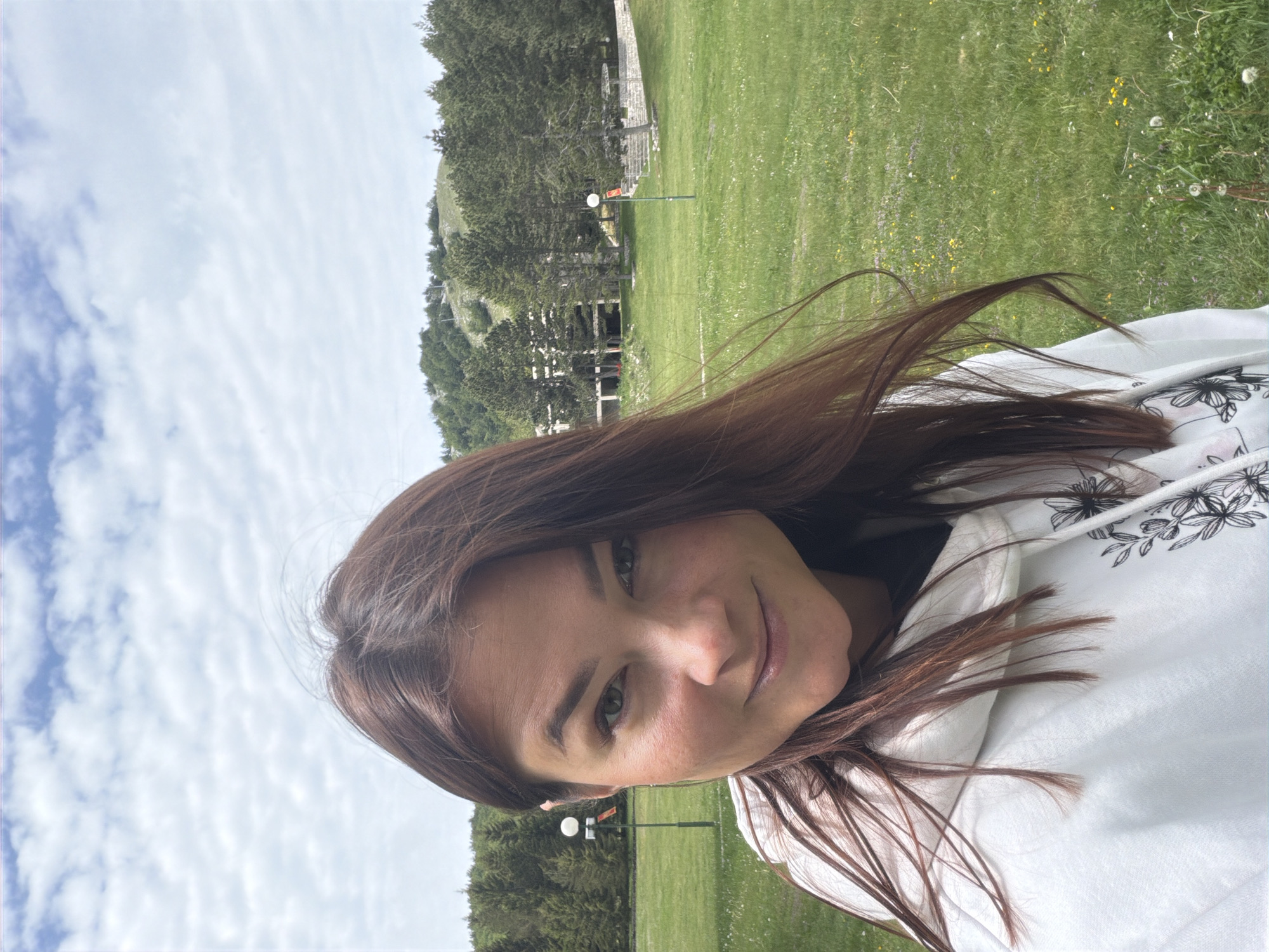 A close-up, portrait-style photograph captures a young woman outdoors against a natural landscape. The woman, positioned in the foreground, has long, dark brown hair that flows around her face and shoulders, appearing slightly wind-swept. She wears a white hooded sweatshirt adorned with black floral patterns and smiles gently towards the camera.
The background reveals an expansive natural scene. The upper portion is dominated by a bright, partly cloudy sky featuring long, thin cloud formations. Below the sky, a vibrant green grassy field, speckled with small yellow and white wildflowers, extends across the middle and lower right parts of the image. A dense line of dark green conifer trees runs horizontally through the mid-ground, partially concealing a hill or mountain. Amongst the trees, hints of human infrastructure are visible, including what appears to be a concrete staircase or structure, a white globe lamp on a pole, and a pole with a small red sign. The setting suggests a mountainous or hilly recreational area, possibly a resort, consistent with Mali Bostur, Montenegro. The photo appears to be taken during the daytime under partially cloudy skies, conveying a serene and natural atmosphere.