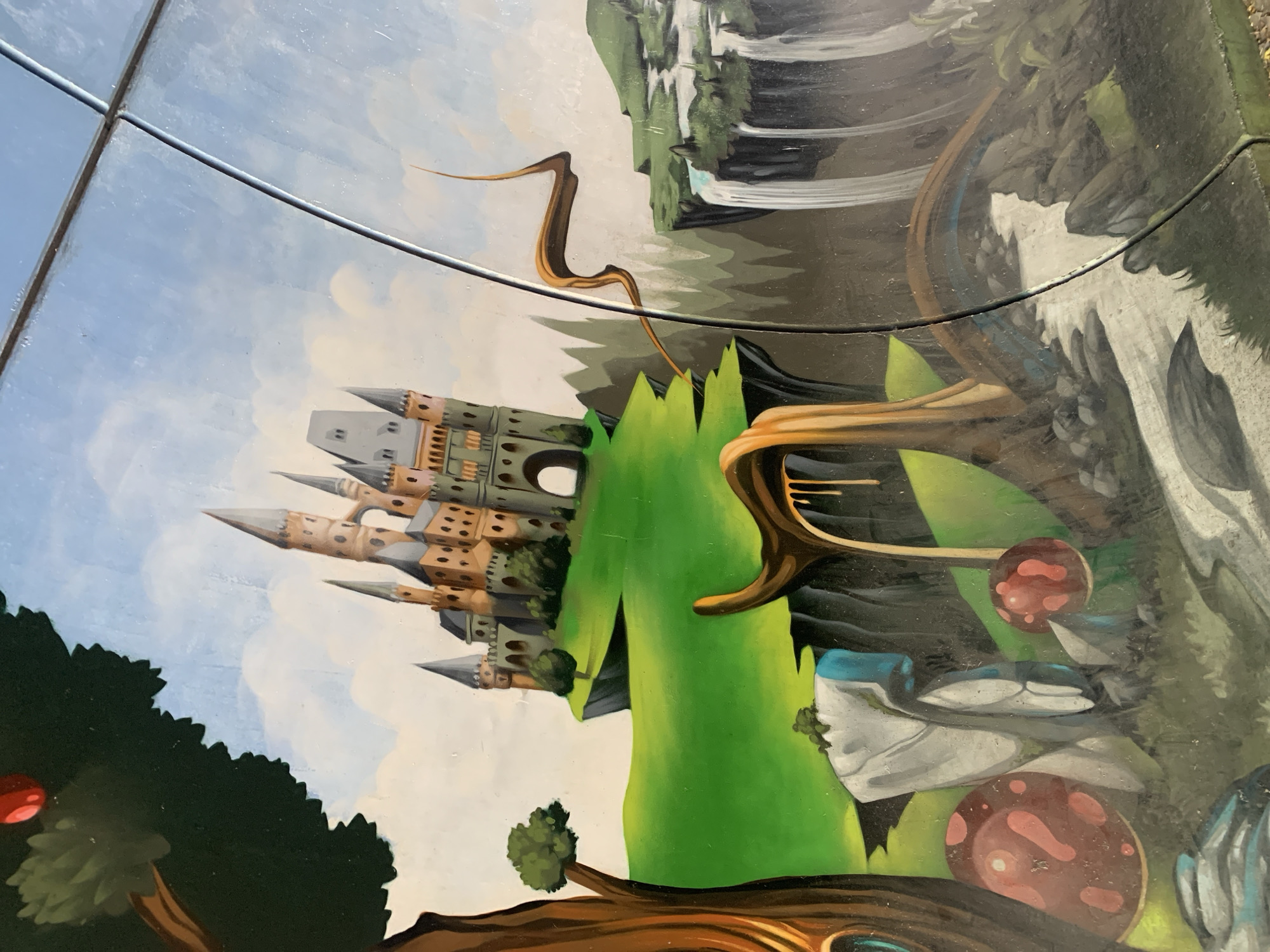 This is a close-up, distorted, overhead photograph of a mural painted on a glass surface. The mural depicts a fantastical, dreamlike landscape. Dominating the left side is a multi-turreted, tan and grey castle with pointed spires and square windows, seemingly built from blocks or dice. It sits on a green, undulating hill. To the right of the castle, a large, vibrant green, organic shape curves upwards, resembling a stylized plant or mountain. Further to the right, a waterfall cascades down a rocky cliff face into a dark, reflective pool. Other elements of the surreal landscape include abstract, wood-like structures, large, spotted red mushrooms, and rock formations. A dark green, jagged-topped tree with a red orb resembling a fruit or planet is visible in the lower left. The sky is painted with soft blues and whites, suggesting a daytime scene. The mural appears to be viewed through a clear, possibly reflective material, with horizontal lines, possibly the edges of glass panels or wires, bisecting the image. The overall impression is one of an otherworldly, imaginative realm. The location context is Odivelas, Portugal.