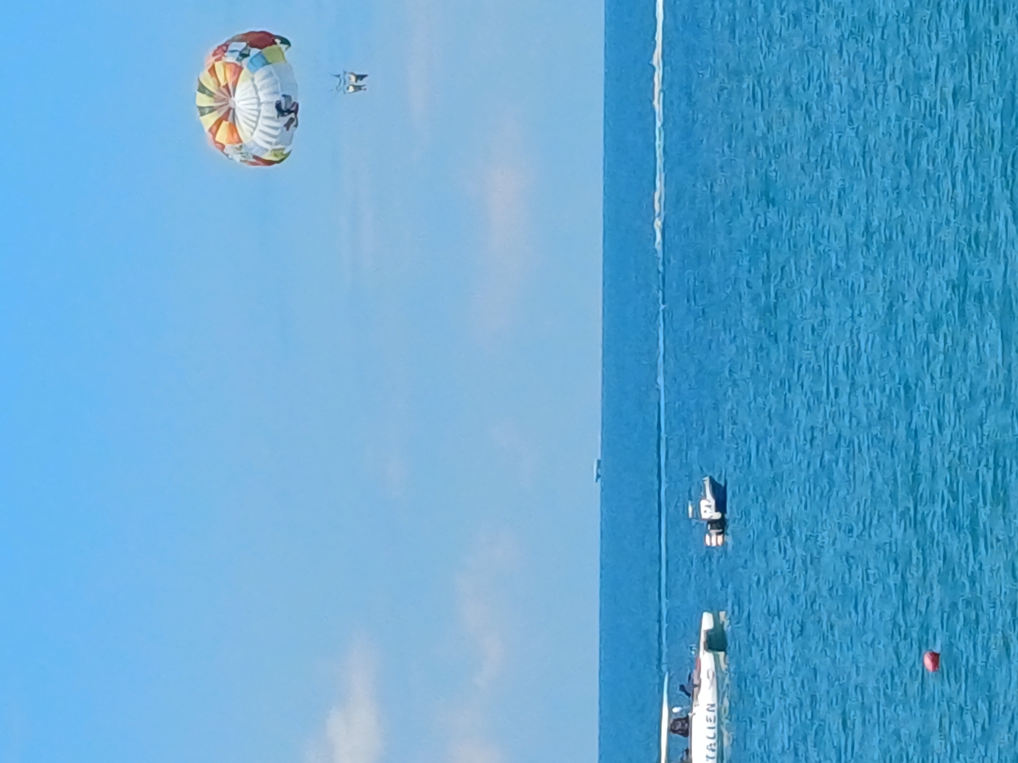 A bright, sunny day with a clear blue sky over a calm, blue sea. In the distance, a colorful parasail with two people attached floats high in the air. Closer to the shore, two boats are visible. One is a white speed boat with the word "ITALIEN" written on its side, and the other is a smaller blue and white boat. A red buoy floats in the foreground. The scene depicts a typical recreational day at the seaside, possibly in Sochi, Russia, given the context.
