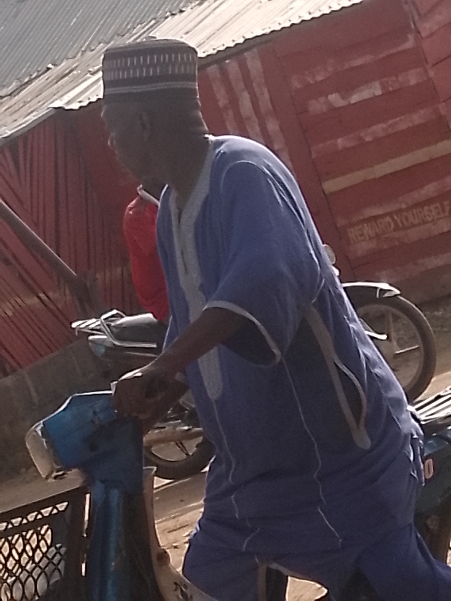 A man wearing a dark-colored hat and a blue robe is riding a light-blue motorcycle in what appears to be an urban environment. Behind him, a person in a red shirt is riding another motorcycle. A red structure with the phrase "REWARD YOURSELF" is in the background. The location is Unknown, Unknown.