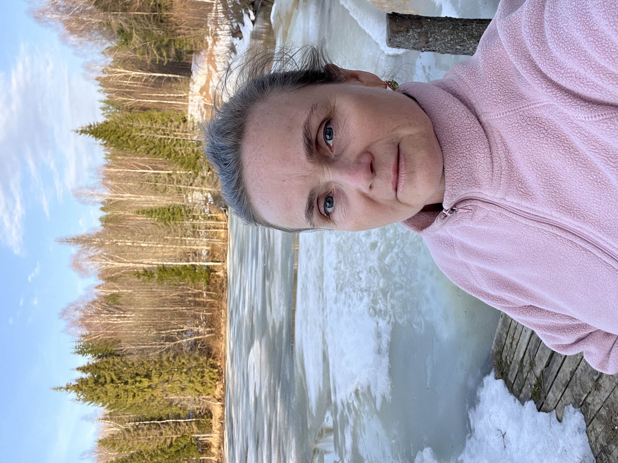 A woman in a pink fleece jacket stands on a wooden structure, likely a dock or platform, overlooking a partially frozen body of water. Ice formations are visible on the water's surface. In the background, there is a forest with bare trees and some evergreen trees. The sky is blue with scattered clouds. The image appears to be taken during late winter or early spring. The location is Zhigalki, Russia.