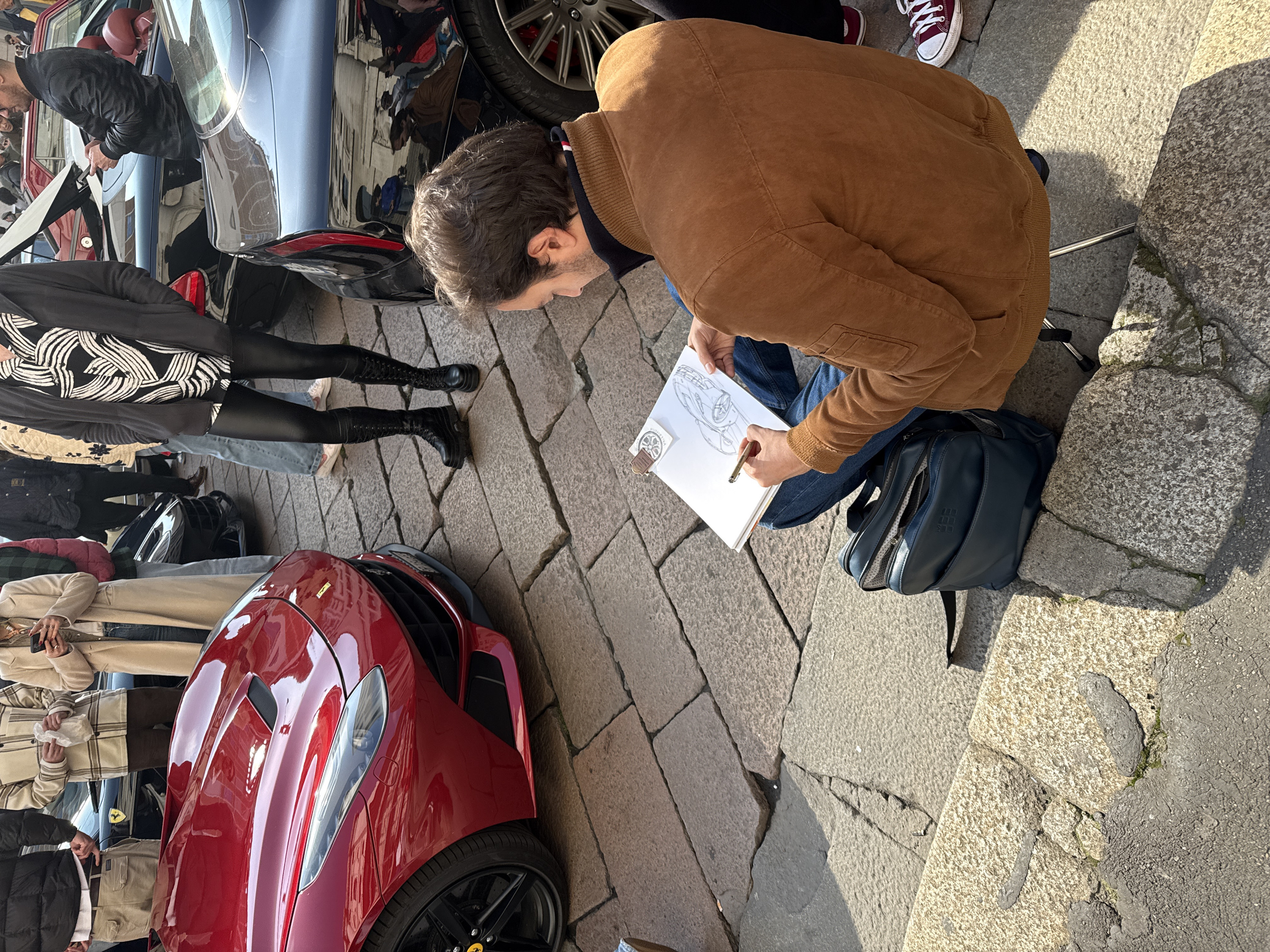 The scene depicts an outdoor gathering of individuals viewing luxury sports cars on a cobblestone surface in the city of Milan, Italy.
In the foreground, a male individual is seated on a portable stool, facing right, meticulously sketching a sports car on a white pad with a gold-colored pen. He wears a brown suede-like jacket over a dark blue collar with red and white stripe accents, and blue jeans. An open dark blue Moleskine backpack rests on the cobblestones to his left.
A highly polished, dark red Ferrari F12berlinetta occupies the left-midground, angled slightly towards the right. Its front fascia, left headlight, engine hood vents, and the front left wheel with black multi-spoke rims and a yellow Ferrari emblem are distinctly visible. The car's surface reflects the surrounding architecture and sky.
Behind the red Ferrari, a group of people observes the vehicles. This group includes a person in a black puffer jacket and tan cargo pants, adjusting their belt; another in a beige plaid shacket holding a crumpled white bag; and a female in a long beige coat and blue jeans, looking at a smartphone. Further back and to the right, another person in a black shirt with white patterns and black pleather-like pants, wearing black platform boots, stands near a black sports car.
A black luxury sports car is partially visible behind the artist and to the right of the red Ferrari, showing its rear left wheel with red brake calipers. In the background, a dark blue convertible with a red interior is parked with its trunk open. Another black vehicle featuring a yellow Ferrari badge on its fender is partially visible behind the foreground crowd.
The ground is paved with large, irregular cobblestones in varying shades of gray and beige, with visible wear, cracks, and small patches of moss in some interstices. Strong natural sunlight illuminates the scene, casting distinct shadows.