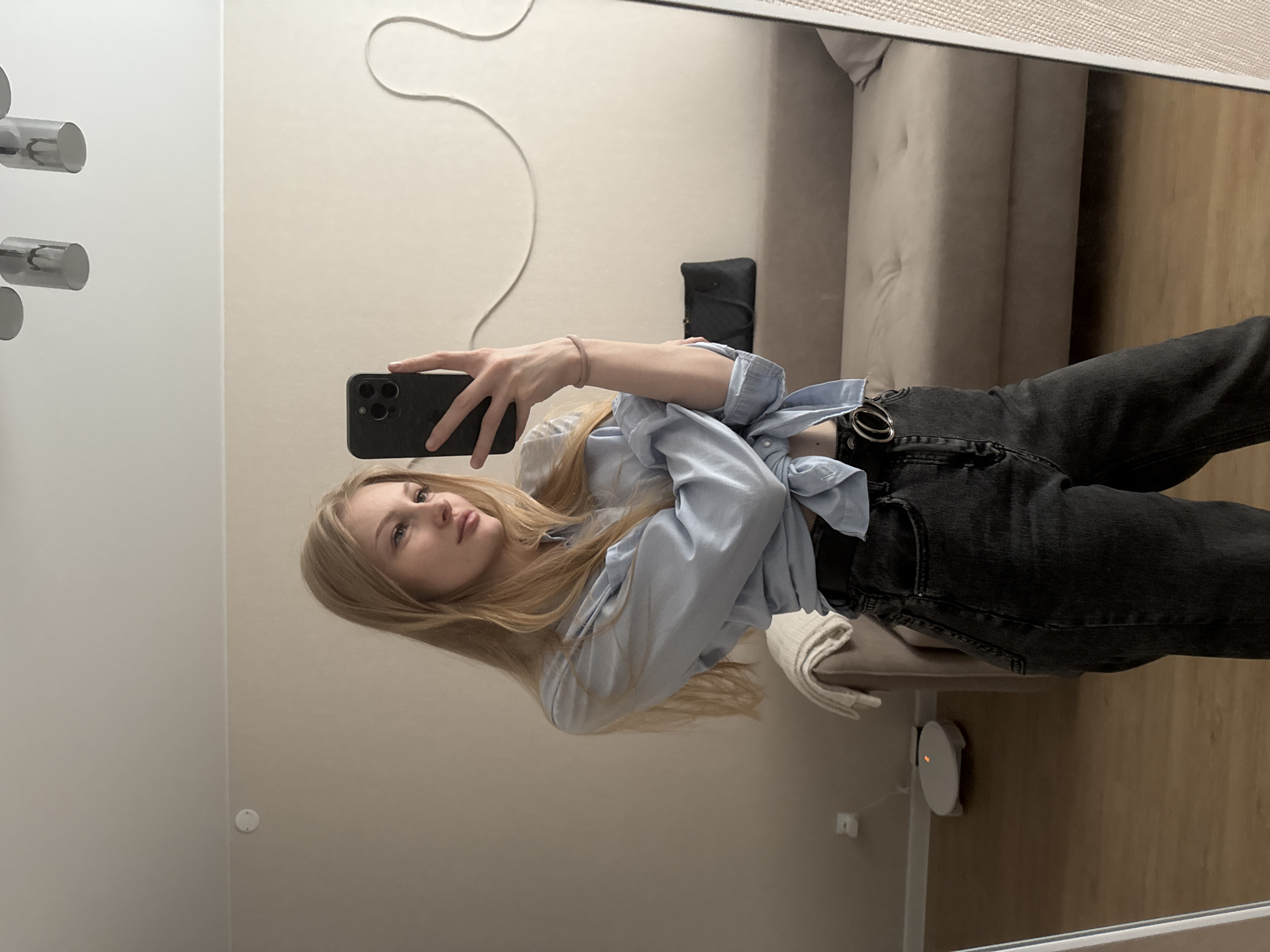 A young woman with blonde hair takes a mirror selfie in a room. She is wearing a light blue button-down shirt tied at the waist, exposing her midriff, and dark wash jeans with a silver belt. The room has light beige walls, a wooden floor, and a beige sofa. A robotic vacuum cleaner is visible on the floor in the background. The image appears to be taken indoors, likely in a residential setting. The metadata of the image indicates it was taken in Perm, Russia.