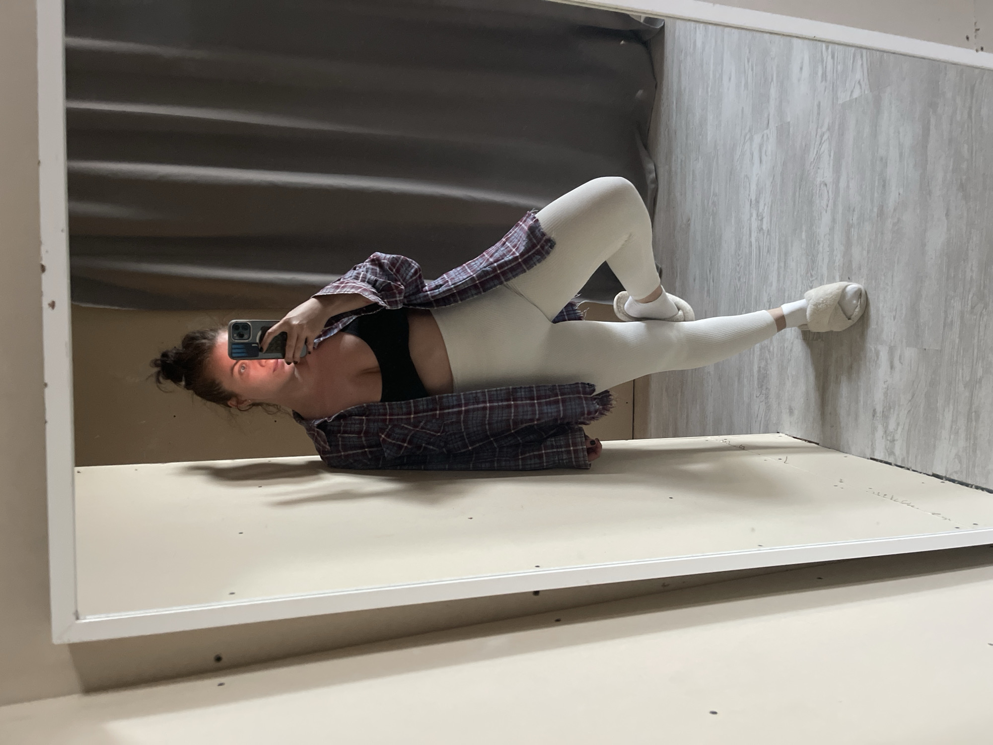 A woman takes a photo of herself in a mirror. She is wearing leggings, a sports bra, and a plaid shirt. She is wearing slippers. She is in an indoor space with a curtain and a wooden-look floor. There is no indication the location is Мосметаллоконструкция Онт, Russia.