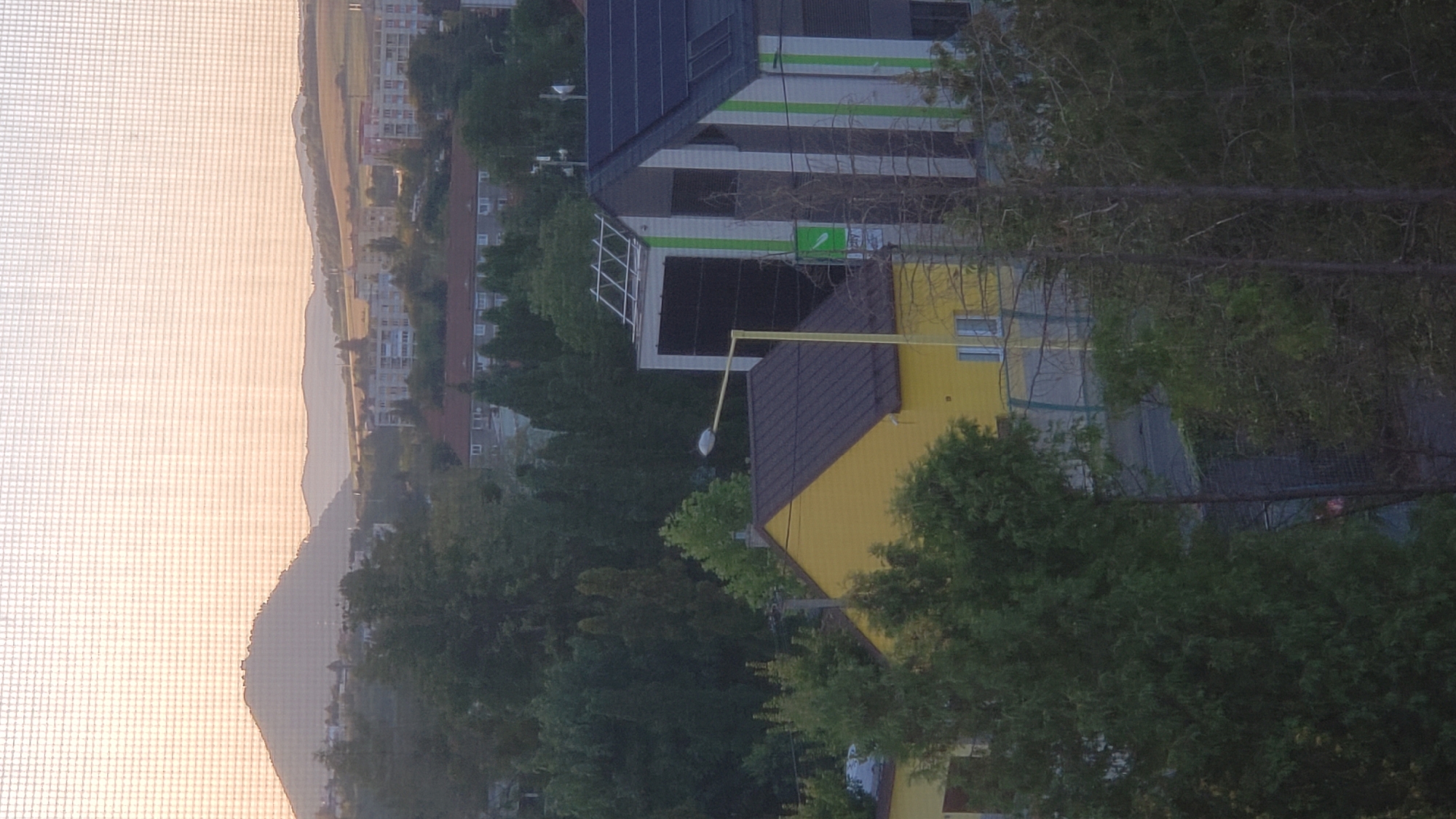 The image is an outdoor shot of a cityscape, likely taken from a window as a screen mesh is visible across the top. The scene depicts a residential area with a mix of building types, including a bright yellow house with a brown roof in the foreground. Adjacent to it is a modern building with a gray and white striped facade, topped with solar panels. Trees of various sizes and types are scattered throughout the scene, with some tall evergreen trees prominent on the left. In the background, there are apartment buildings and hills under a hazy, possibly sunset sky. The overall impression is a suburban or semi-urban environment. The specific location within Prešov, Slovakia cannot be determined from the image alone. There are no people or discernible actions captured in the frame.