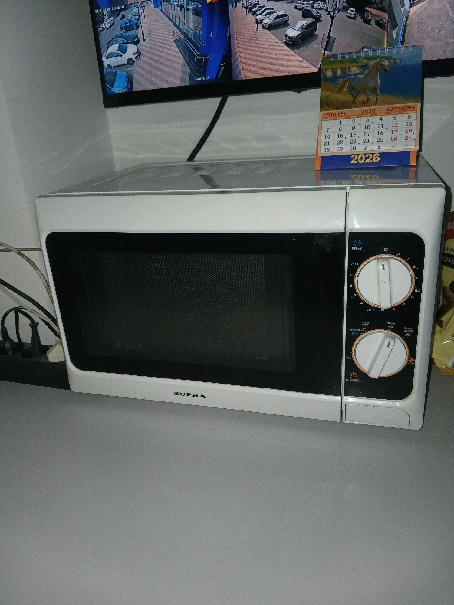 A white Supra brand microwave is positioned on a gray surface in what appears to be a domestic or office setting. The microwave has a black door with a mesh pattern and control knobs on the right side. The control panel includes two circular dials, one for time and another for power level. The time dial is set to approximately 30 minutes, and the power dial is set to a medium level, indicated by a symbol of three wavy lines. Above the microwave, a digital screen displays a CCTV feed of a parking lot with several cars. To the right of the screen, a desk calendar for September 2026 is visible, featuring an image of a horse. The calendar is open and shows the days of the week and dates. A power strip with several plugs and cables is located to the left of the microwave. The background is a plain gray wall.