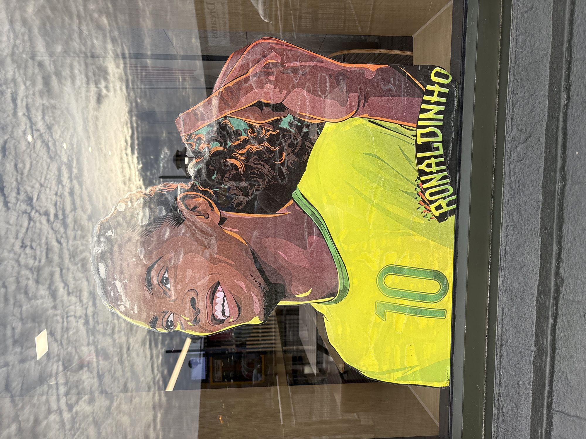 A stylized illustration of Ronaldinho, a male soccer player, is affixed to a glass window. He is depicted wearing a bright yellow jersey with the number "10" in green on the front. His name, "RONALDINHO," is written in a green, stylized font below the jersey, accompanied by orange stars. The illustration is vibrant, with bold outlines and a dynamic color palette. Reflections of a cloudy sky and an indoor environment, possibly a restaurant with tables and chairs, are visible on the glass, overlaying the artwork. The lower portion of the image shows the textured surface of the window frame and a gray brick wall.