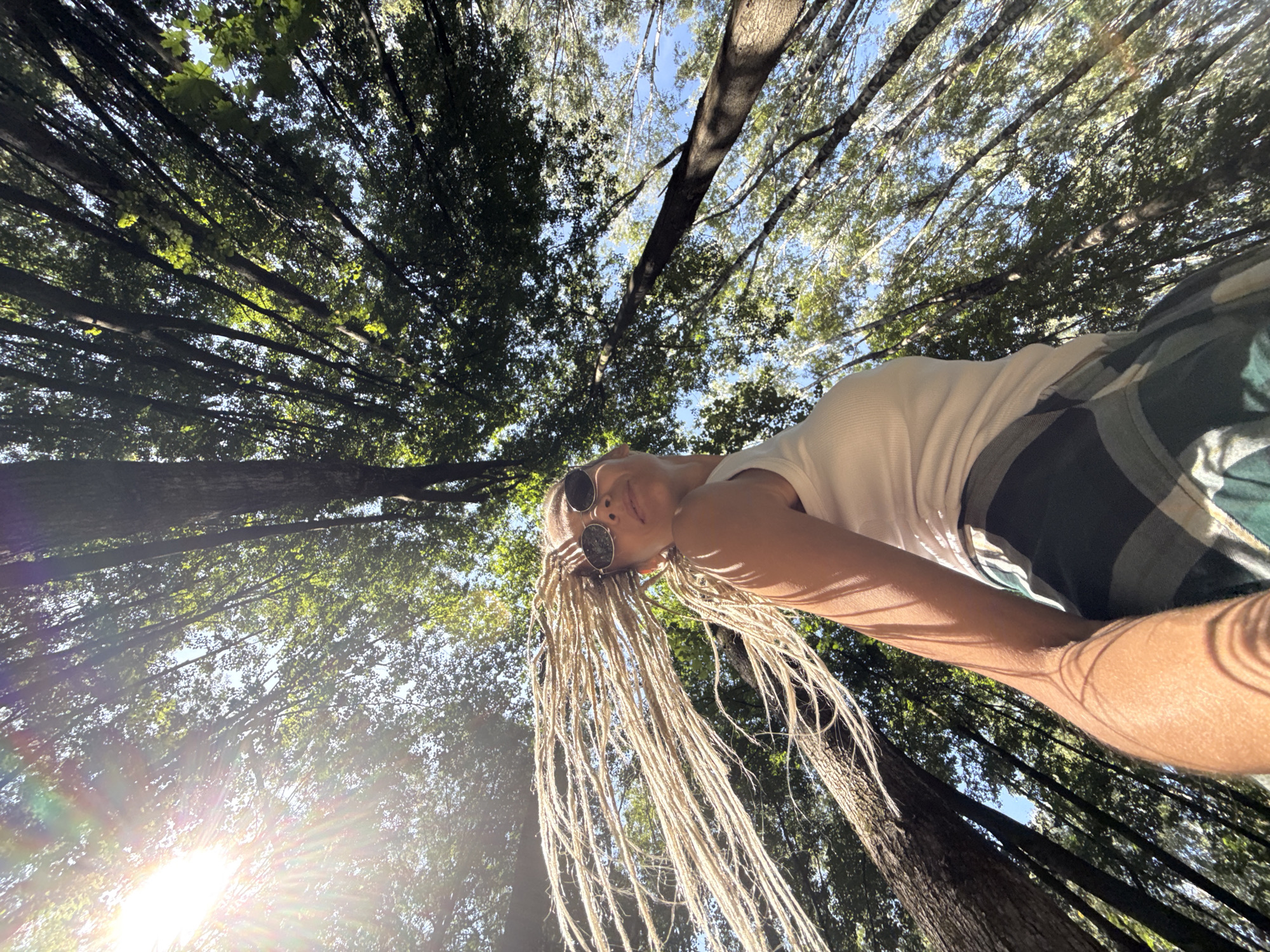 A low-angle, wide shot captures a woman with long, light-colored dreadlocks in a forest in Rusanovo, Russia. She is wearing a white tank top and plaid shorts and is looking up at the camera. Her dreadlocks are being blown by the wind, creating a dynamic effect. The sun is shining brightly through the trees, casting lens flares and dappled light on the scene. The trees are tall and dense, with green leaves and dark bark. The sky is a clear blue. The woman's arm and part of her torso are visible in the foreground, with shadows cast by the tree branches.
