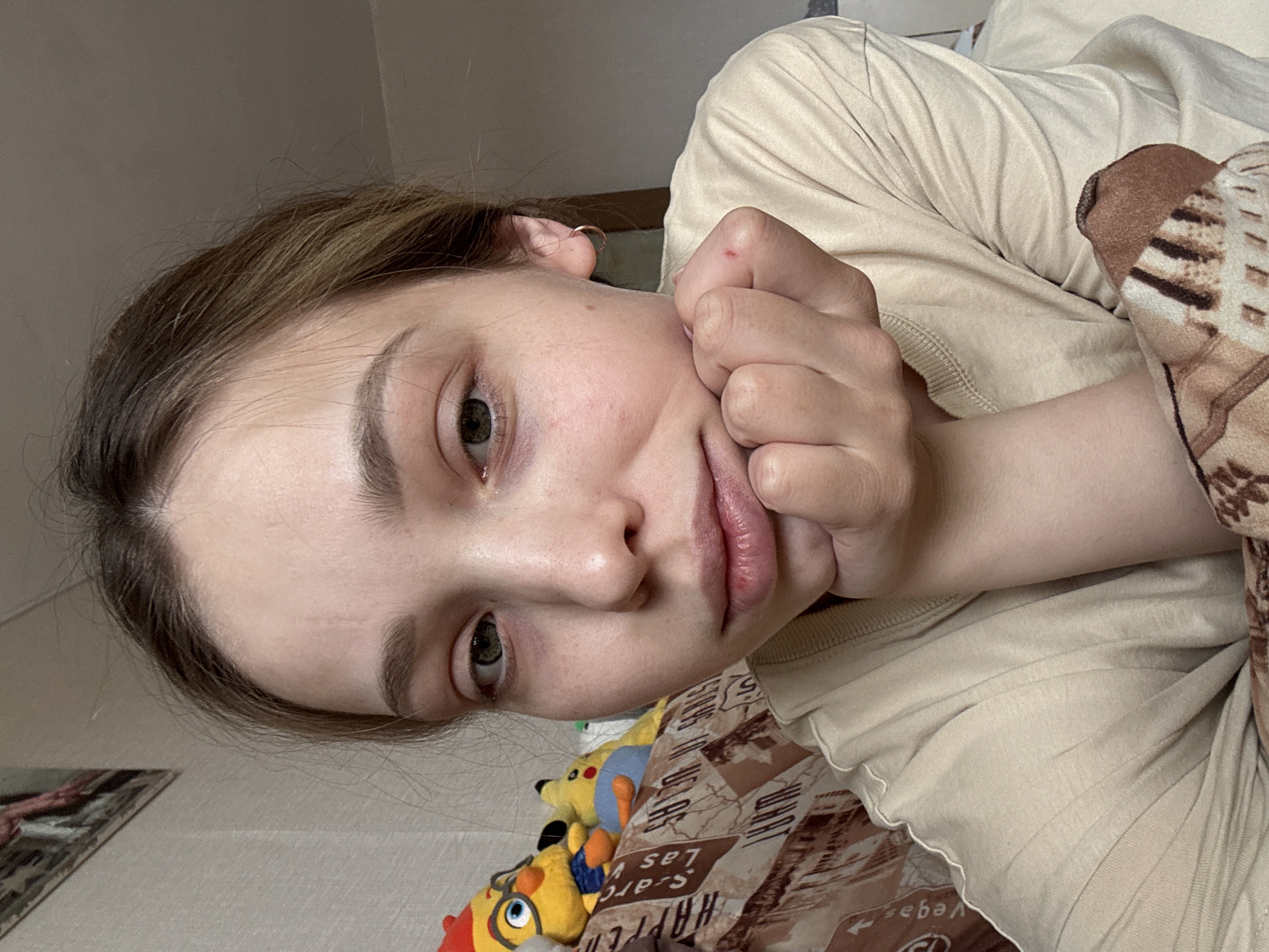 This image captures a close-up of a young adult female, approximately late teens to early twenties, in an indoor residential setting located in Chaikovsky, Russia. The subject has light brown hair, green eyes, and a fair complexion, exhibiting minor blemishes. She wears a light beige or cream-colored long-sleeved ribbed top and a small golden hoop earring in her right ear. Her left hand is positioned under her chin, with fingers curled, and a small red mark or scab is visible on the knuckle of her left ring finger. Her gaze is directed towards the camera.
In the background, behind her left side, several plush toys are visible on a patterned surface, likely a bedspread or blanket. These toys include a yellow cartoon bird wearing glasses and blue shorts, and a partially obscured yellow figure resembling Pikachu. The bedspread features a brown and beige pattern with partially discernible text such as "Las Vegas" and "Route 66." The wall behind the subject's head on the left is a light, vertically textured surface, with a portion of a framed picture visible in the upper corner. The upper right portion of the background consists of a plain, light brown or beige wall. The lighting appears soft and ambient, illuminating the subject's face evenly.