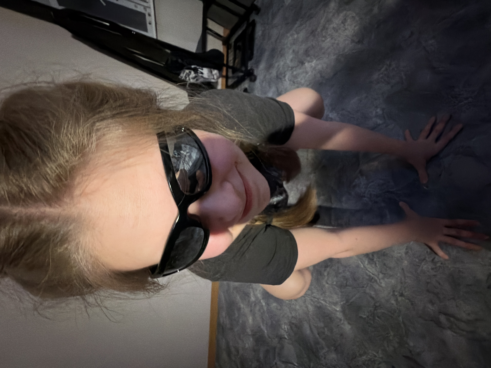 A young person with blonde hair is kneeling on a gray textured surface that appears to be a floor. They are wearing dark sunglasses and a dark-colored, short-sleeved top. Their hands are extended forward, touching the floor. In the background, there is furniture including a black rack or shelf and a white object. The location in the city of Perm, Russia is not able to be ascertained from the picture provided.