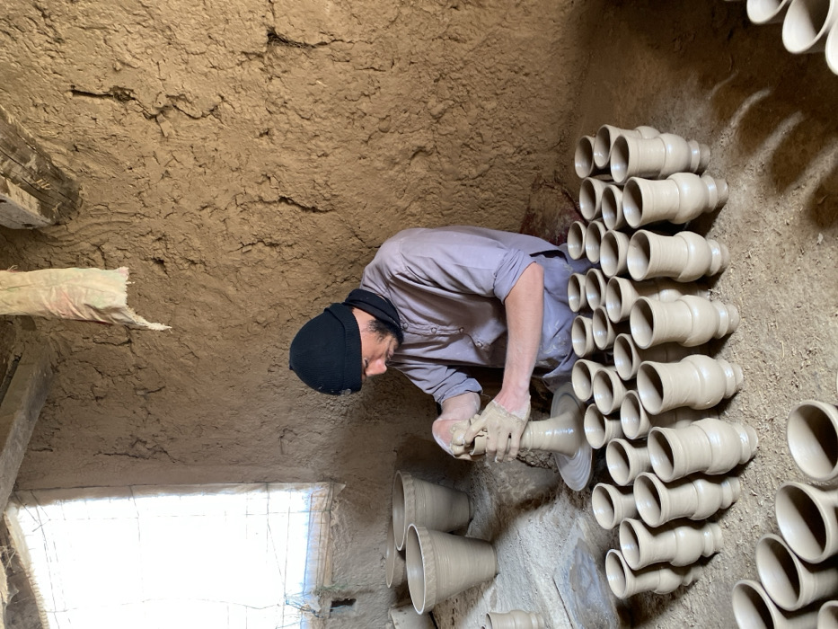The image depicts an indoor pottery workshop in Kandahar, Afghanistan. An adult male, dressed in a black knit beanie, a dark neck warmer, and a light-colored long-sleeved shirt, is seated on the earthen floor. He is actively engaged in shaping a light-colored clay vessel on a rotating pottery wheel with both hands, which are visibly coated in clay.
Numerous unglazed, light brown clay vessels are present throughout the scene. Approximately forty to fifty smaller, footed vases or cups are meticulously arranged in rows on the earthen floor in front of and to the right of the potter. Three larger, unglazed clay planters or pots are visible behind the potter, positioned near a window on the left wall.
The workshop features rough, uneven, mud-plastered walls that show cracks and textures consistent with traditional construction. A rectangular window, covered with a mesh or screen, is on the left wall, admitting bright natural light into the space. The floor is earthen and appears dusty. A light-colored fabric scrap hangs from a wooden beam in the upper right portion of the image.