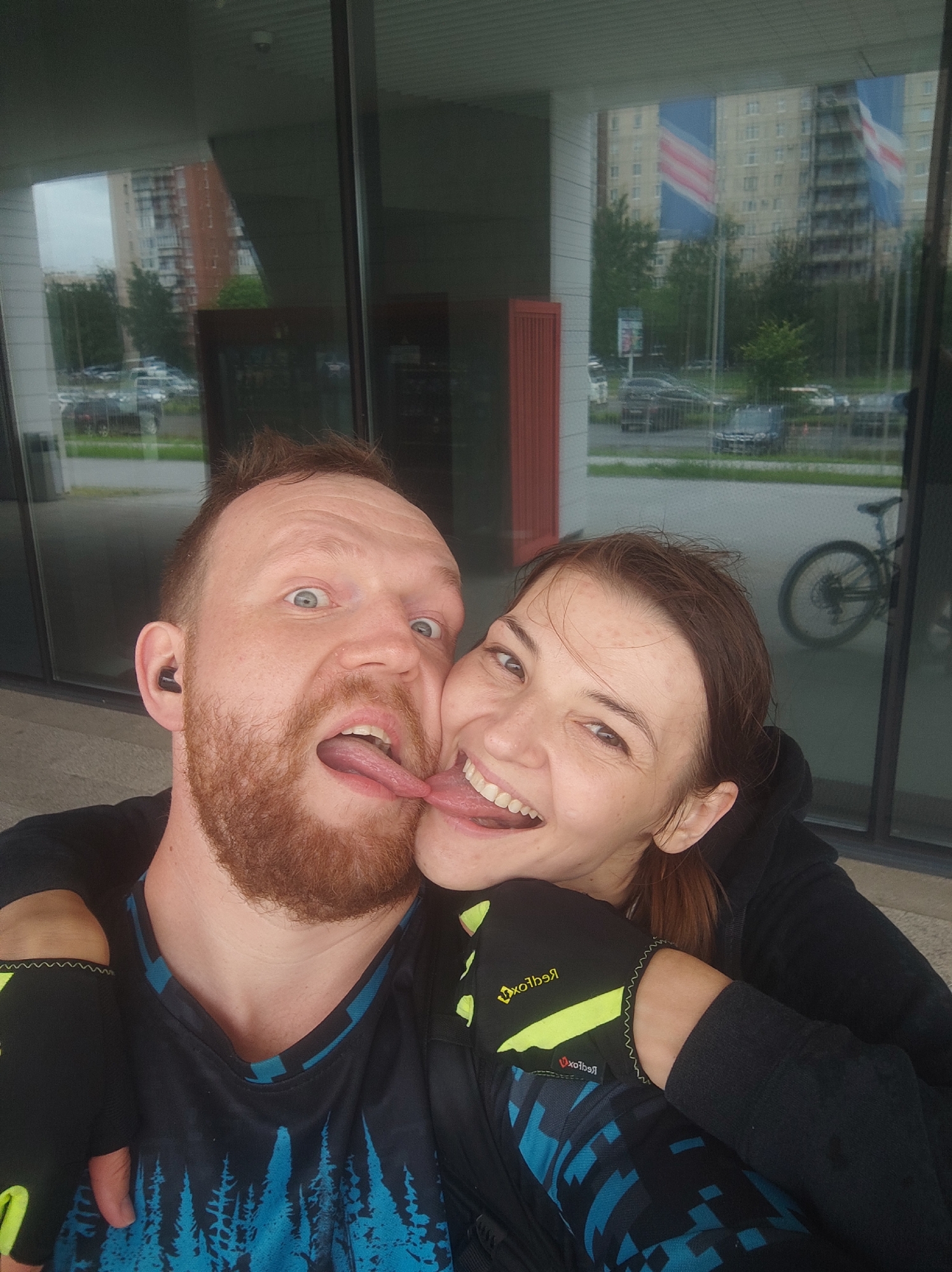 A selfie is taken of a Caucasian man and woman with their tongues out, playfully touching each other. They are outdoors in front of a modern building with large glass windows. The man has a red beard and is wearing a black shirt with a blue and black tree pattern. The woman has brown hair and is wearing a black hoodie. In the background, through the glass, are reflections of apartment buildings, trees, cars, and flags. The environment appears to be an urban setting in Saint Petersburg, Russia, possibly a residential or commercial area, indicated by the architecture and presence of flags. The overall mood is lighthearted and intimate.